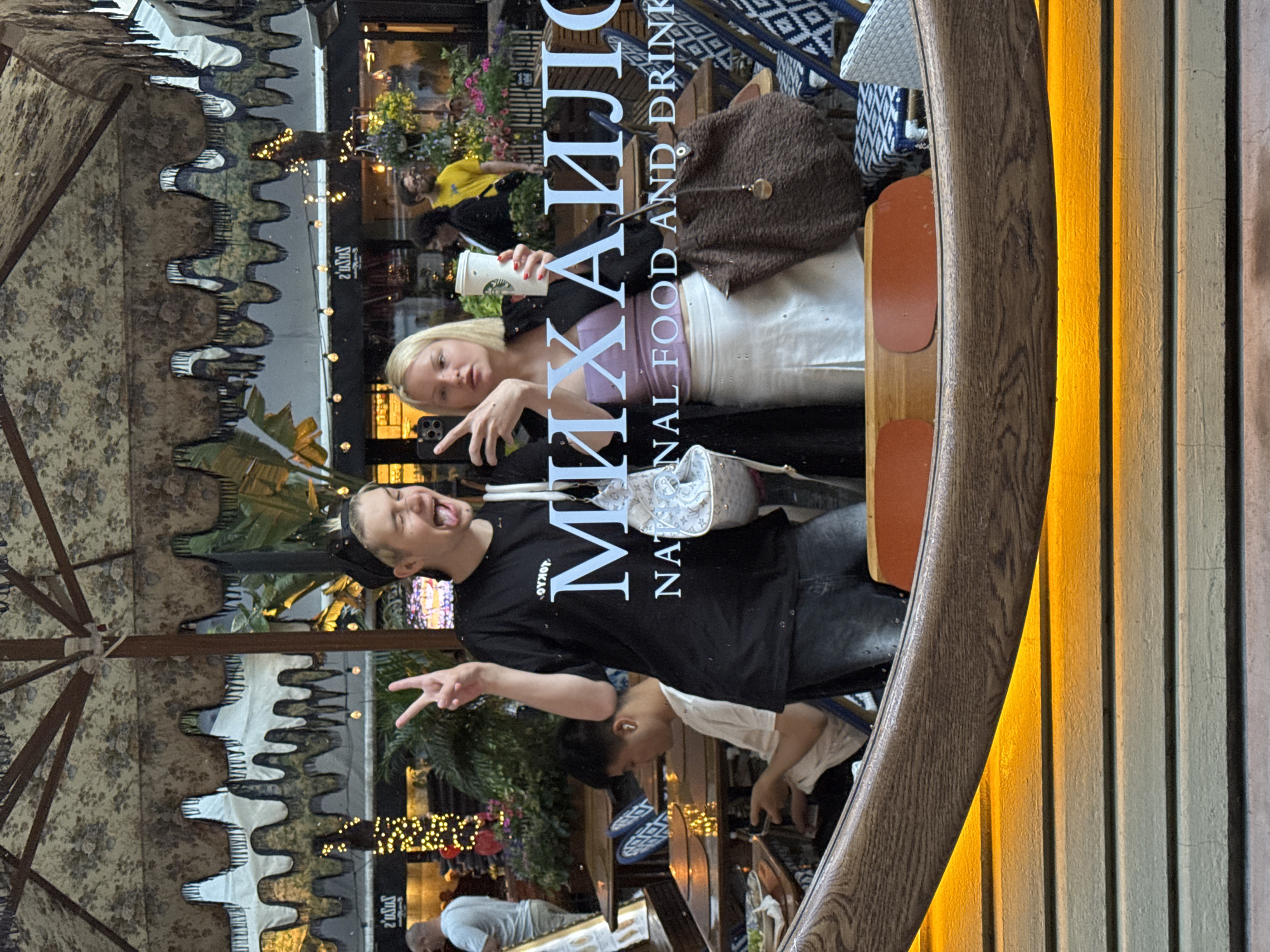 The image shows a mirrored reflection of two people posing for a photo. One person, with blond hair, is holding a phone, likely taking the picture, while holding a cup. The other person is making a peace sign with their hand and sticking out their tongue. The reflected text indicates "MIXA," "NATIONAL FOOD AND DRINK." The surrounding environment suggests an outdoor dining area with umbrellas and lights, likely in a public space. Based on the text, this is likely the "MIXA" restaurant in Belgrade, Serbia.