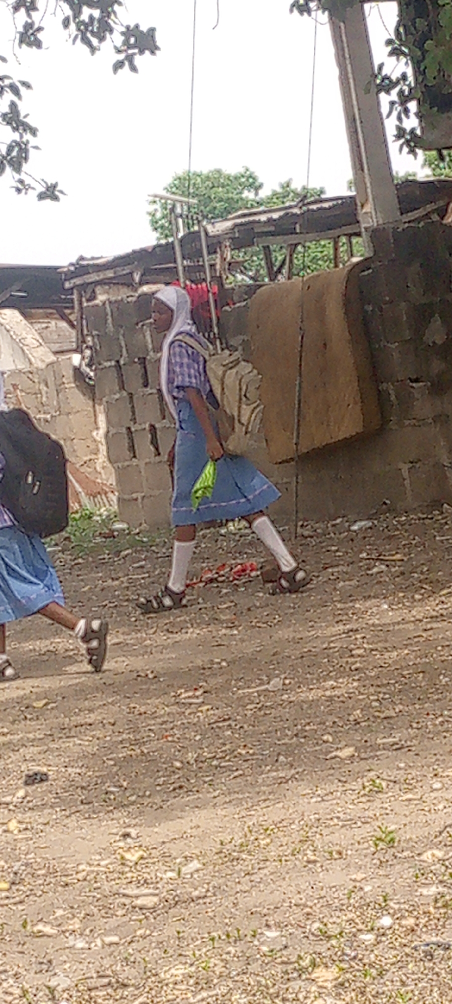 This media file captures two young individuals, likely schoolgirls, walking outdoors on a dusty, unpaved path. The image is taken in Adankolo, Nigeria, and shows a scene under natural daylight.
The primary subject, positioned centrally, is a girl seen in profile, walking from right to left. She wears a light-colored hijab, a plaid shirt (possibly blue and white or purple and white checks), a blue skirt, white socks, and dark sandals. A light-colored backpack is slung over her shoulders. In her right hand, she carries a bunched-up green item, possibly a piece of fabric or a plastic bag. Her expression is neutral as she proceeds along the path.
To the left, partially visible, is another individual, likely also a schoolgirl, wearing a similar blue skirt, white socks, and sandals, with a dark backpack on her back.
The setting appears to be a semi-urban or rural environment. The background features structures made of concrete blocks, some appearing unfinished or in disrepair. A large, brown, possibly fabric-covered board or mattress leans against a concrete block wall behind the central figure. Overhead, tree foliage is visible against a bright, overcast or light-filled sky. The ground is rough and unpaved, covered in dirt, small stones, and sparse vegetation. The overall impression is one of everyday life in a modest neighborhood, with the girls engaged in the routine activity of commuting, likely to or from school. No visible text is present in the image.