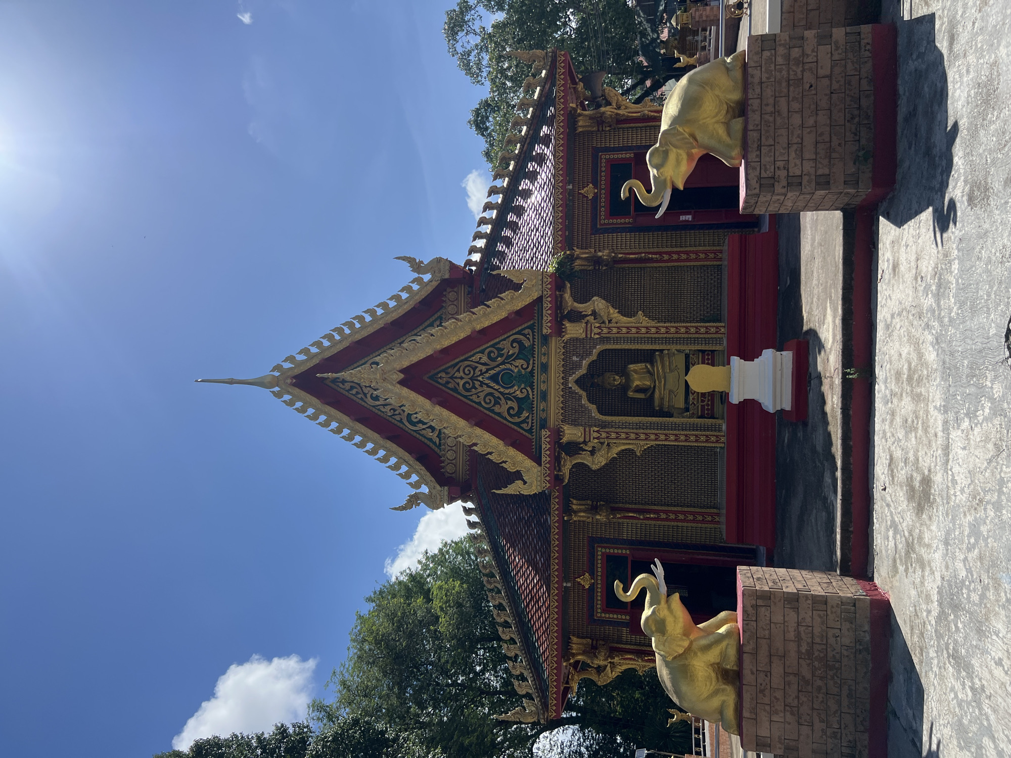 The image shows a Buddhist temple in Thep Krasatti, Thailand, under a bright blue sky with scattered clouds. The temple facade is ornate, featuring a multi-tiered roof with red tiles and gold decorative elements. A statue of the Buddha is visible in a central niche. Flanking the entrance are two large, golden elephant statues seated on brick pedestals. The ground in front of the temple is paved with concrete, and shadows from the elephant statues are cast across it. Trees with lush green foliage surround the temple, suggesting a natural setting within the city.