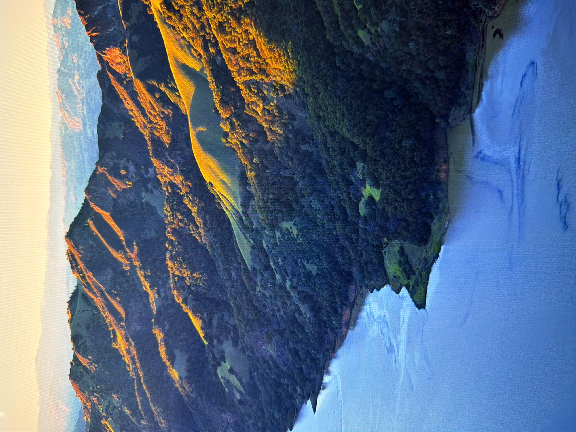 This is an aerial, vertical shot of a landscape at sunrise. The foreground is dominated by a large body of blue water, likely a lake or reservoir, with subtle ripples on its surface. The water meets a dark, tree-covered shoreline that curves into the distance.
Behind the shoreline, a series of rolling hills rise, covered in a mix of dark green trees and lighter green patches that suggest open grassland or meadows. The late morning sun casts long shadows across the slopes, highlighting the contours of the terrain. The light is a warm golden color, illuminating the tops of some hills and the edges of the tree lines.
In the background, more distant mountain ranges are visible, fading into a hazy, pale blue and yellow atmosphere, indicative of the sunrise. The overall impression is one of a serene, natural landscape bathed in the soft light of early morning.
This image is not located in the city of Kyiv, Ukraine. The scene depicted is a natural landscape with hills, forests, and a body of water, which is not characteristic of urban Kyiv.