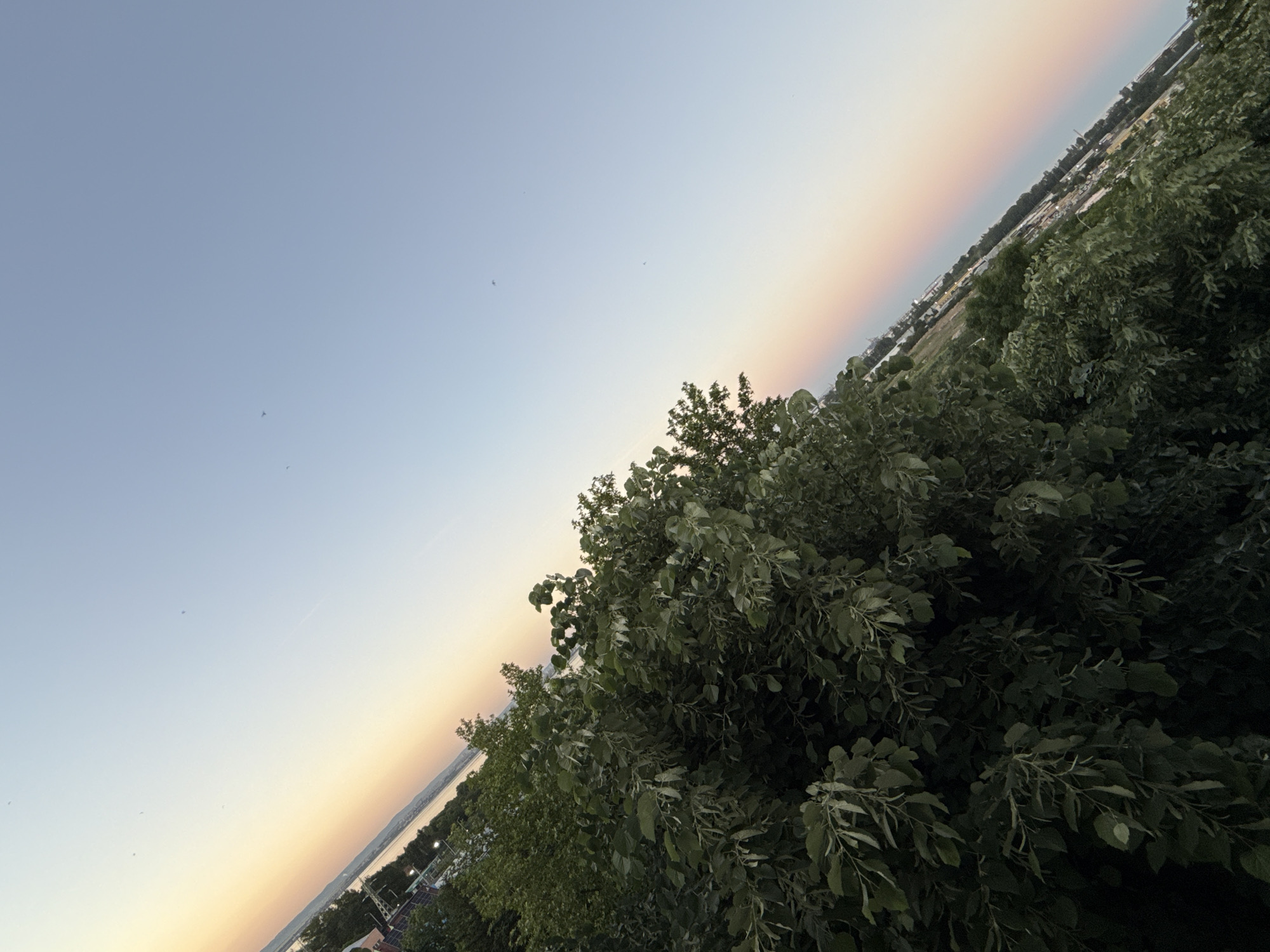 The image shows an elevated view of a landscape. The sky is light blue with a gradient to orange near the horizon. Patches of green foliage, likely trees and bushes, dominate the right side of the image. Buildings and land can be seen in the distance. The picture appears to be tilted to the left. The location is likely Burgas, Bulgaria, given the context of the prompt. Details about specific people, objects, actions, or interactions are not discernible from the image.
