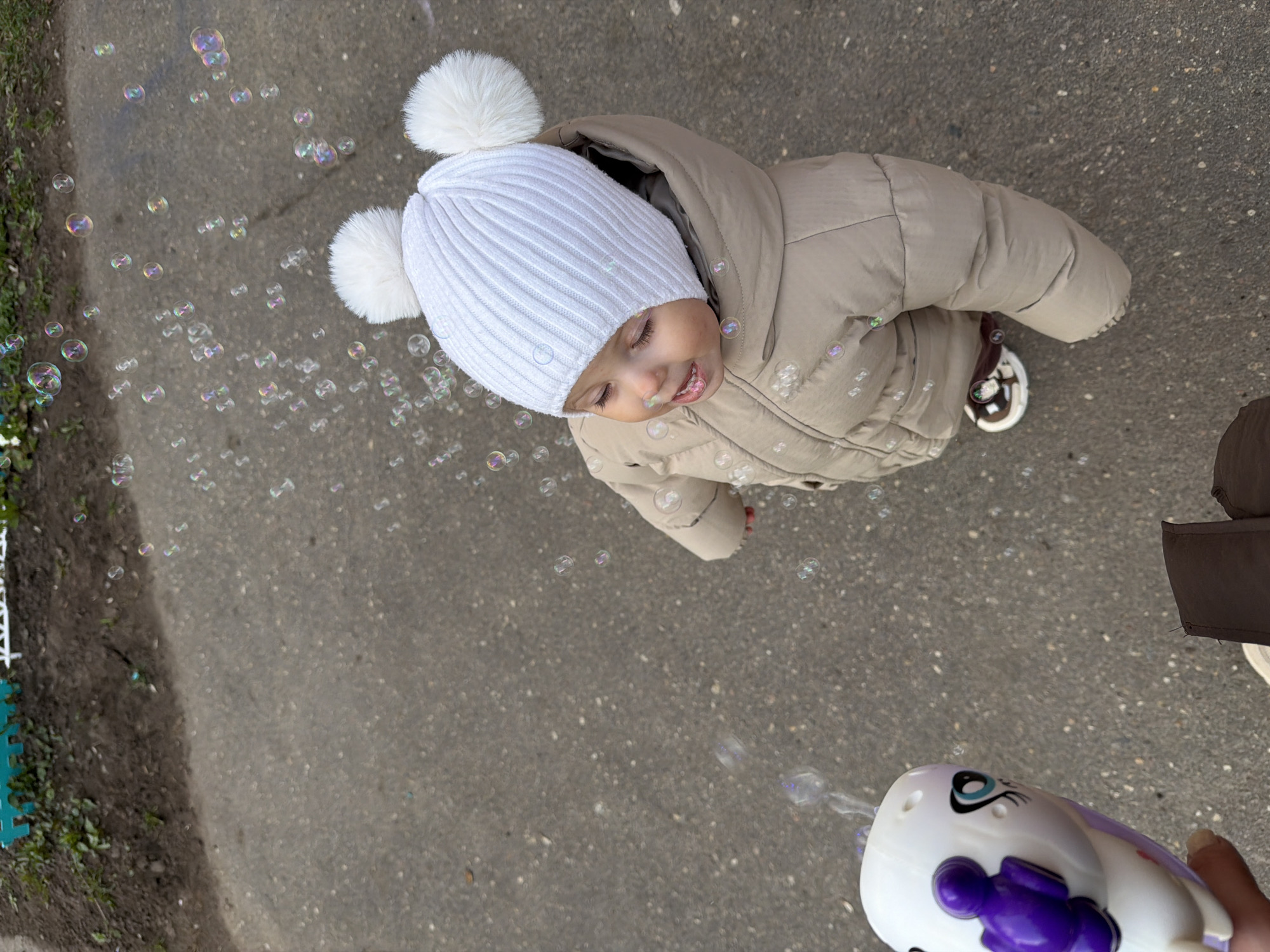 A high-angle shot shows a young child in Reshetnikovo, Russia, dressed in a tan winter coat and white knit hat with pom-poms, looking up and smiling at bubbles floating in the air. The child is standing on a grey asphalt surface, possibly a sidewalk or parking lot. A bubble gun toy, predominantly white with purple accents, is visible in the lower right corner being held by someone partially visible with a brown sleeve. The edge of a green mat or playground surface is visible in the upper left. Bubbles are dispersed around the child and in the area near the bubble gun.