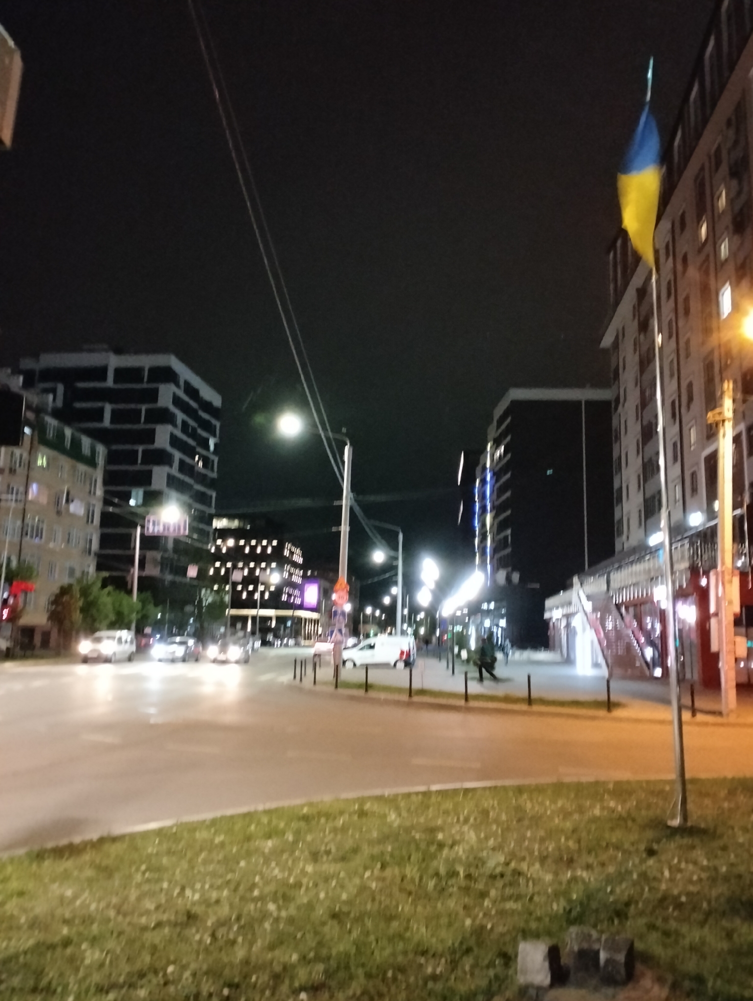 The image depicts a street scene in Ivano-Frankivsk, Ukraine, at night. A Ukrainian flag is prominently displayed on a pole near a light post in the right of the frame. The cityscape features several modern buildings with illuminated windows and decorative lighting. Cars are visible on the road, and a pedestrian appears to be walking on the sidewalk in the distance. There is a pedestrian bridge in the background, and a grassy area is visible in the foreground. The sky is dark, and streetlights illuminate the scene.