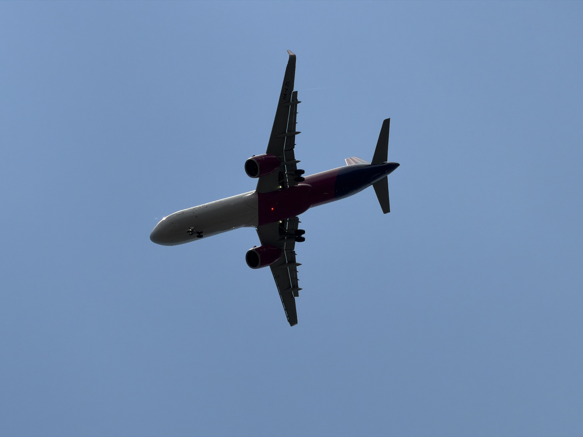 An airplane is pictured from below, against a clear blue sky. The airplane is in flight, with its landing gear extended, suggesting it is either taking off or landing. The underside of the airplane is visible, revealing its engines, wings, and fuselage. The aircraft's tail is colored in shades of dark blue and pink. The registration number "HA-L2L" is visible on the right wing. The image does not contain any information about the city of Burgas, Bulgaria, or any people.