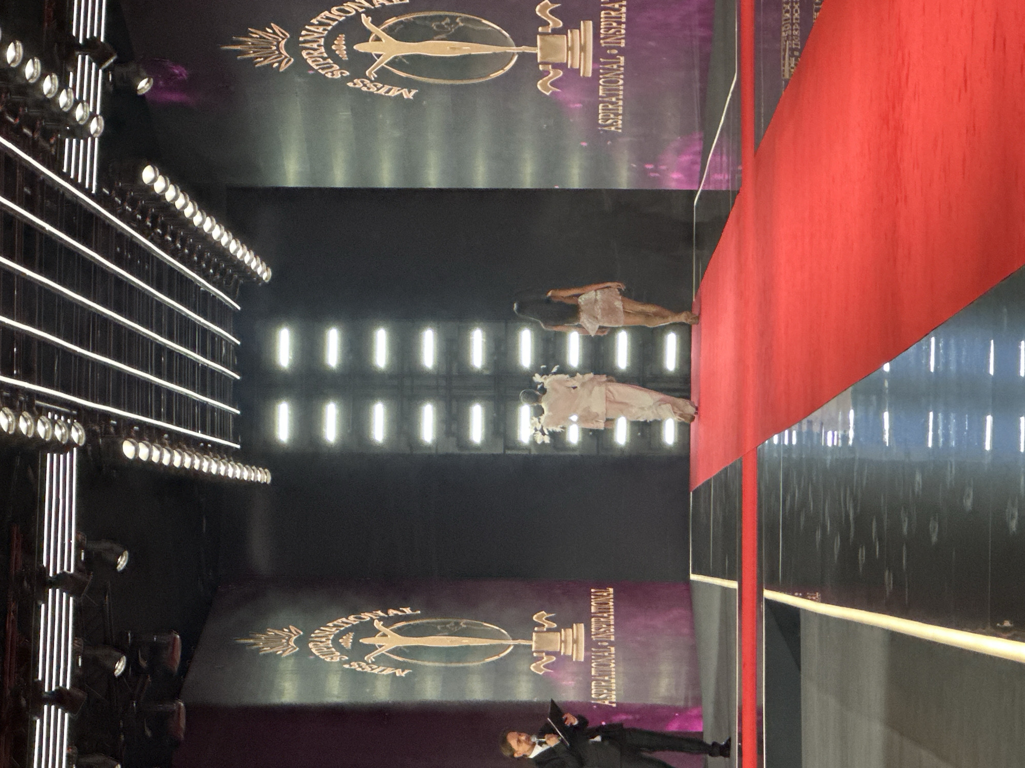 The image captures a formal event, possibly a beauty pageant or awards ceremony, taking place on a red carpet runway. The setting appears to be an indoor venue with a high ceiling featuring intricate lighting structures. On the left side of the runway, a man in a dark suit is holding a tablet and appears to be addressing the audience or participants. He is standing on a dark grey surface adjacent to the red carpet.
The backdrop of the venue is dominated by large screens or walls displaying the "MISS SUPRANATIONAL" logo, accompanied by the words "ASPIRATIONAL - INSPIRATIONAL." These logos are prominently featured on both the left and right sides of the stage, creating a grand visual effect. The screens exhibit a gradient of purple and pink hues.
Two individuals are walking away from the camera down the red carpet. The person on the left is wearing a light-colored, flowing outfit, possibly a suit or a long dress, adorned with feathered elements on the shoulders. The person on the right is wearing a short, sparkly dress. Both individuals are walking towards the center of the stage, which is not fully visible in the frame.
The floor of the runway is reflective, with white horizontal lights running along the edges. The overall atmosphere suggests a professional and glamorous event. The provided crops confirm the presence of the "MISS SUPRANATIONAL" branding and the overall staging of the event.
There is no indication in the image that this event is located in Nowy Sącz, Poland.