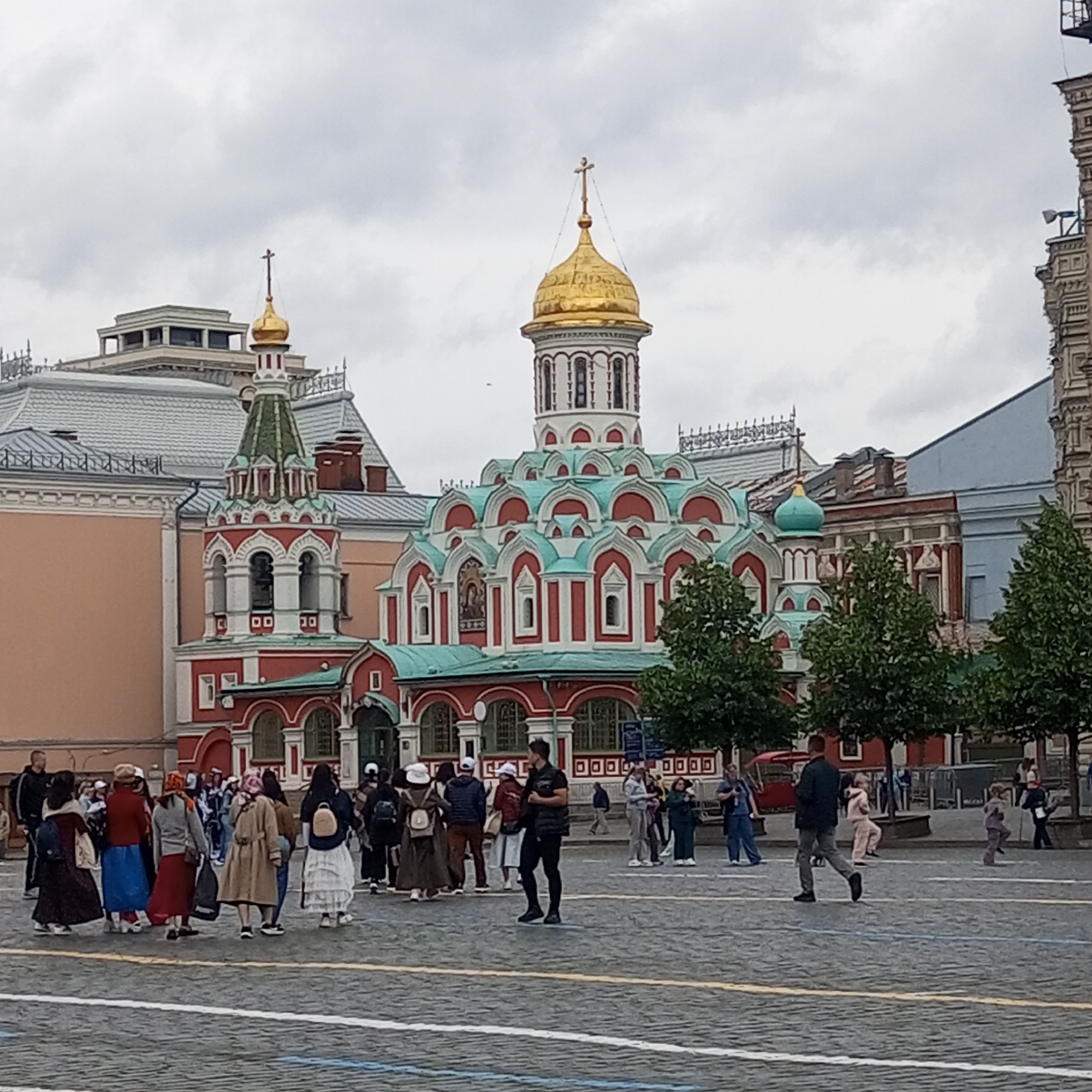 In a paved plaza, a crowd of people stands and walks in front of a red and white building with ornate green domes and a golden cupola topped with a cross. The building appears to be a church or cathedral. The sky is overcast and grey. Several trees line the right side of the scene. The people are dressed in casual clothing, and some are wearing hats. The ground is paved with cobblestones, and white lines are visible on the pavement, possibly indicating pedestrian crossings. A dark-colored vehicle is partially visible on the far left.