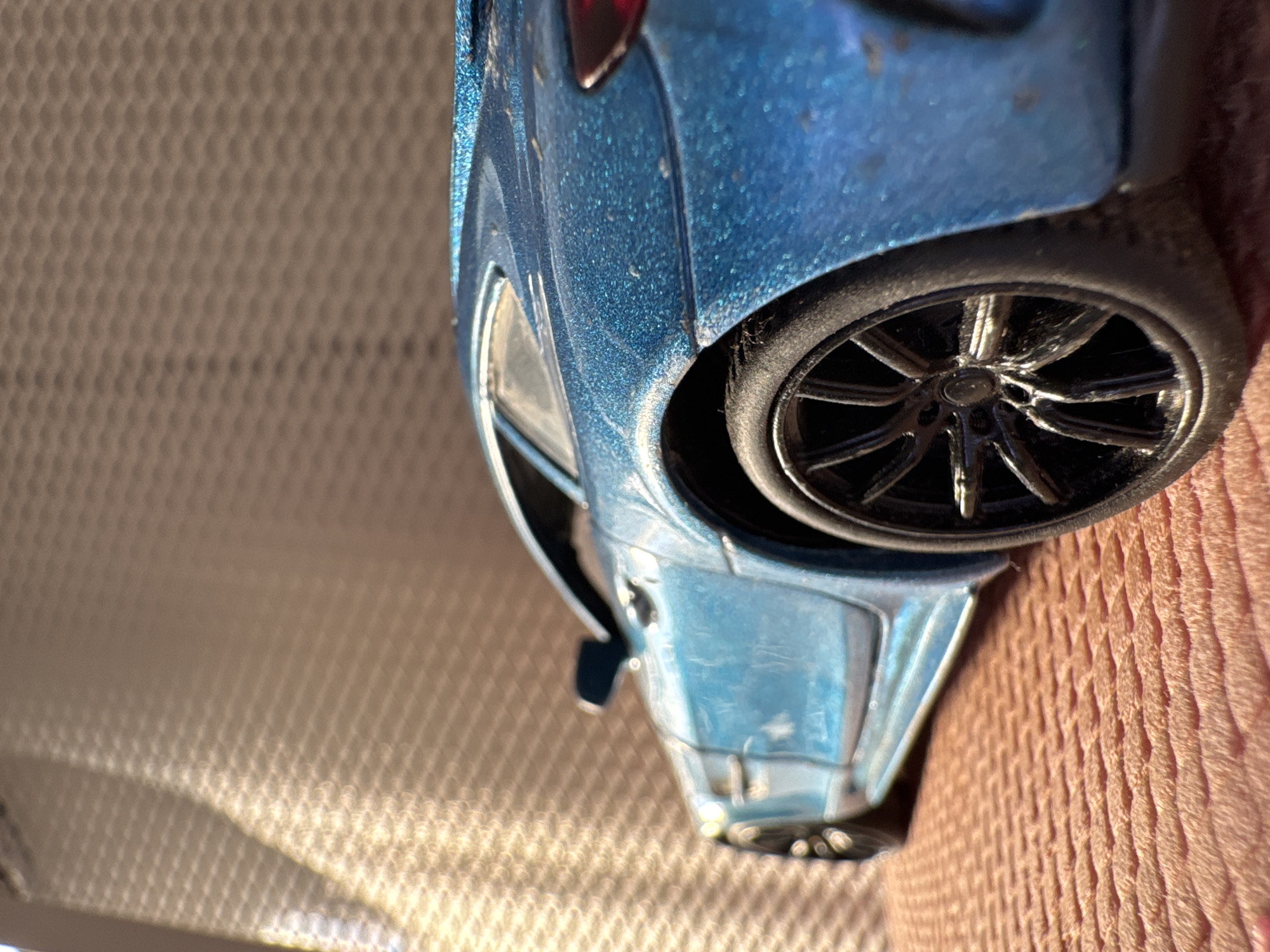 A close-up, rotated view of a blue toy car with a black wheel sitting on a light brown fabric surface. The car's details, including the wheel spokes and body contours, are visible. The background consists of two different textures: a light tan, honeycomb-patterned surface on the left and the brown fabric on the right. The location in Novo-Nikol'skoe, Russia is not discernible from the image. There is no mention of people, subjects, actions, or interactions in the provided image.
