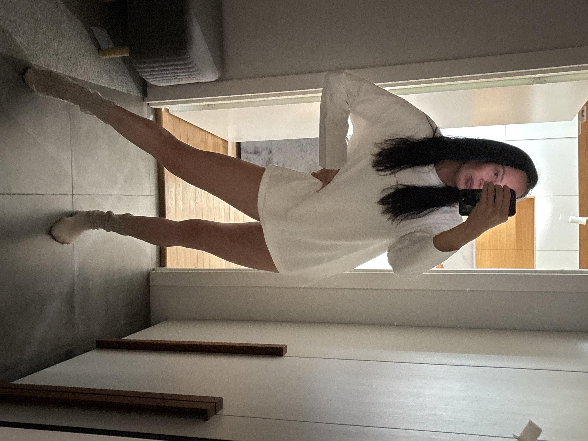 A woman stands in front of a mirror, taking a selfie with her phone. She is wearing a long-sleeved white shirt and socks. The room appears to be a modern bathroom with light-colored walls and wooden accents. Behind her, through a doorway, is a glimpse of a room with a gray couch and a gray floor. There is a light source coming from the bathroom and the room behind it. The location is implied to be in Chaikovsky, Russia.