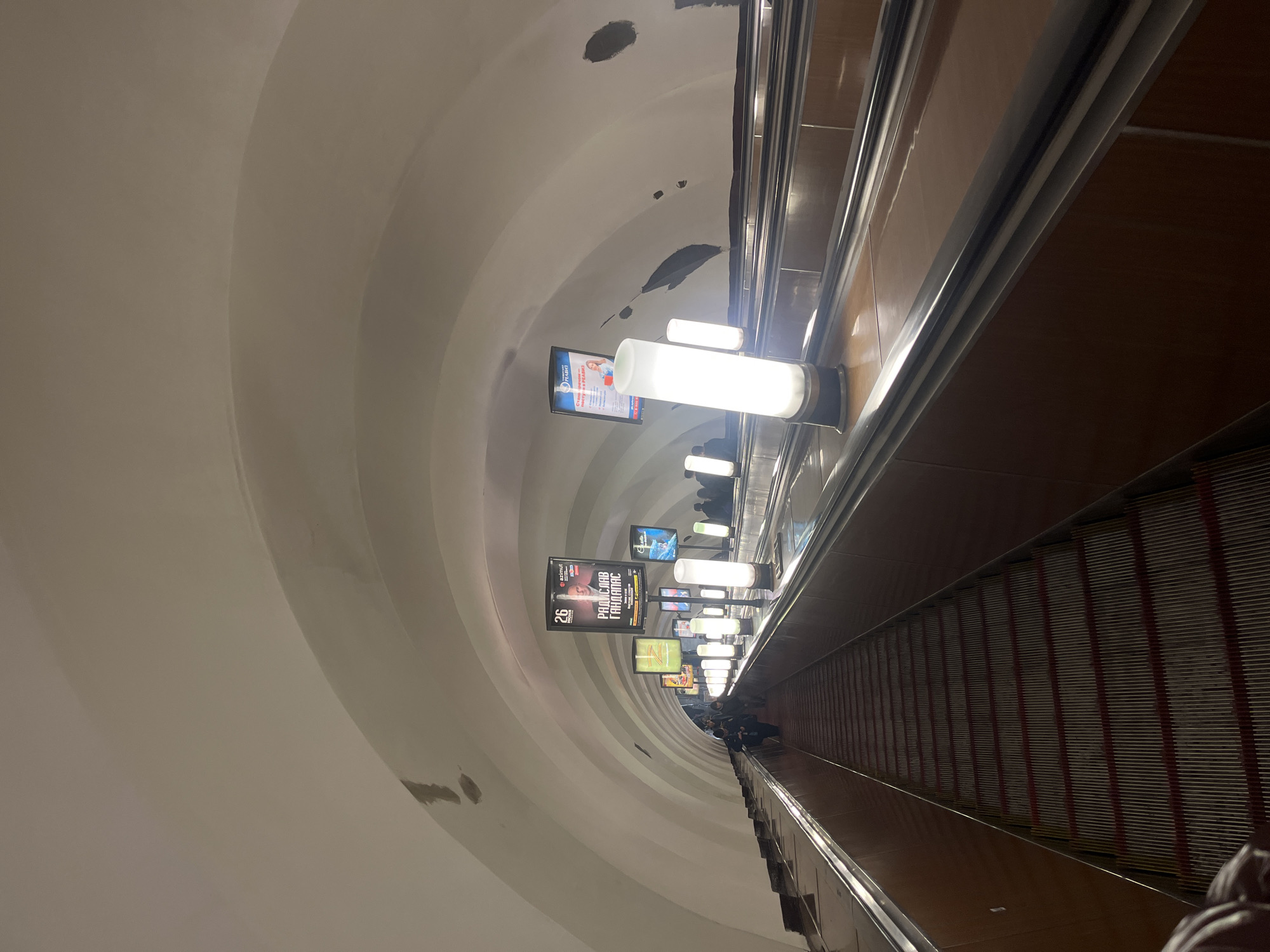 The image depicts a long escalator in a Saint Petersburg, Russia metro station. The escalator is flanked by advertisements and lights. The ceiling is arched and painted white. People are present at the bottom of the escalator. The wall on the right side is paneled in a dark color.