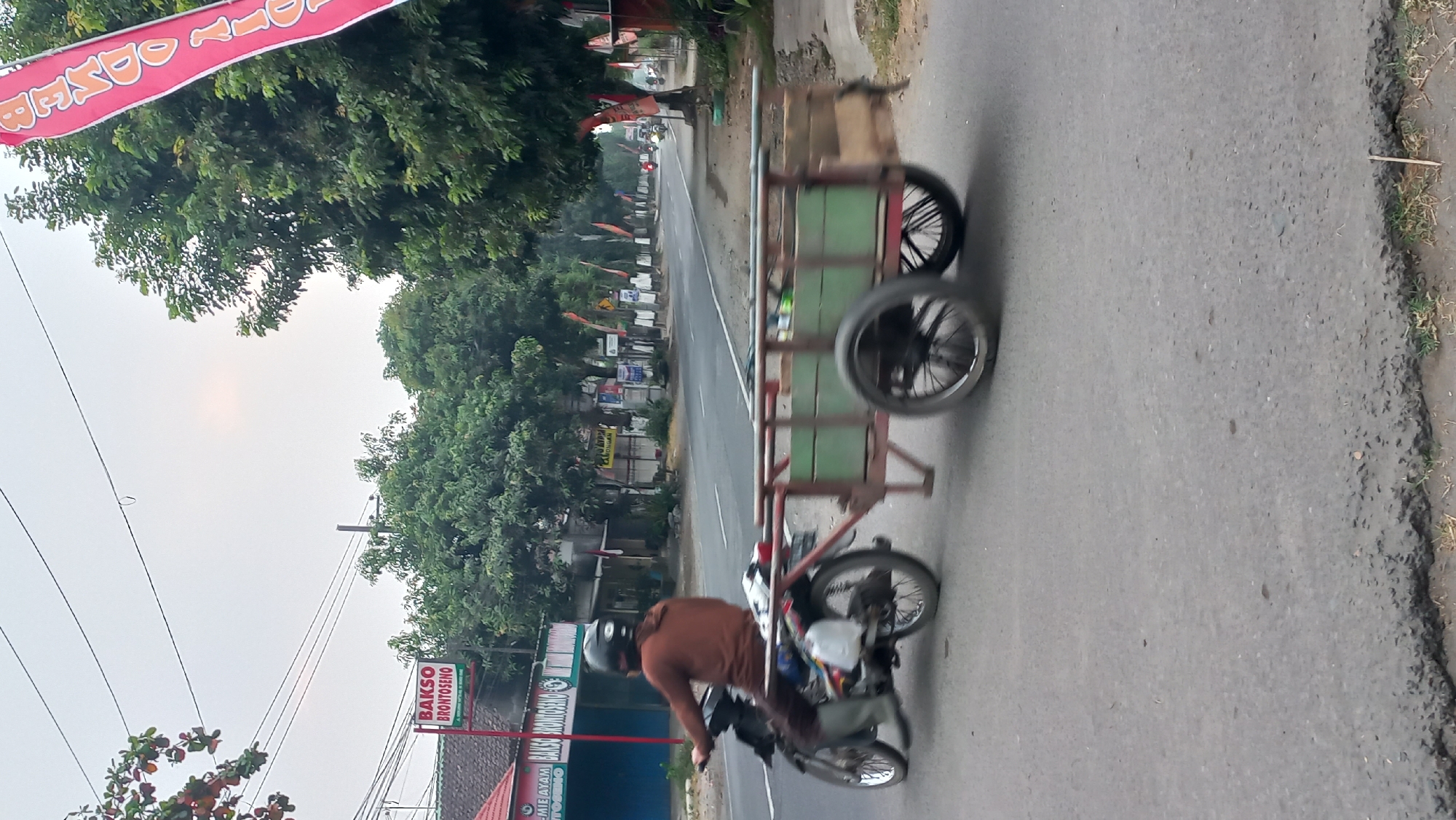 In Pare, Indonesia, on a paved road, a person is riding a motorcycle with a two-wheeled trailer attached. The motorcycle rider is wearing a brown long-sleeved shirt, dark pants, and a black helmet. The trailer is green with a wooden frame and appears to be carrying various items. The road is flanked by trees and buildings, including a sign that reads "BAKSO BRONTOSENO." The sky is overcast, suggesting it might be morning or late afternoon. The image is captured from a side angle, with motion blur indicating movement.