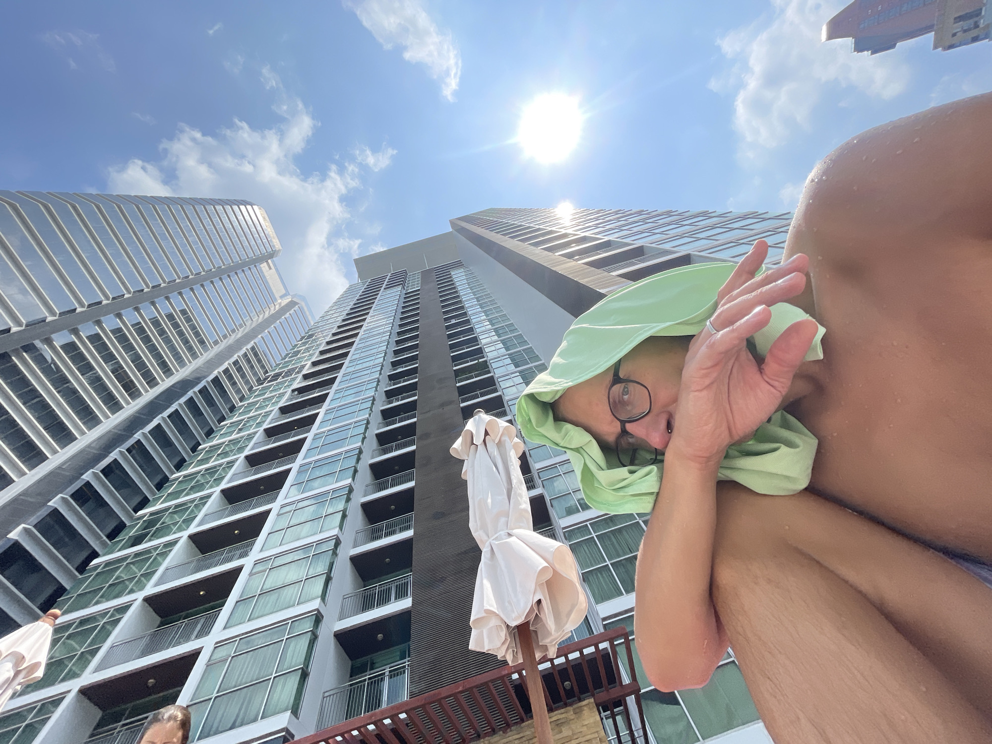 The image shows a person, likely male, wearing eyeglasses and a light green garment or towel covering their head, shielding their face from the sun. The individual is seated, with their arm raised partially obscuring their face. Water droplets are visible on their skin, suggesting they may have just been swimming.
The background reveals a sunny sky with some clouds. Tall modern buildings dominate the cityscape, with balconies visible on the facade of the middle building. An off-white sun umbrella is placed in between the camera and the central building.
Based on the architectural style and general appearance, the location is likely Bangkok, Thailand, a city known for its modern skyscrapers.