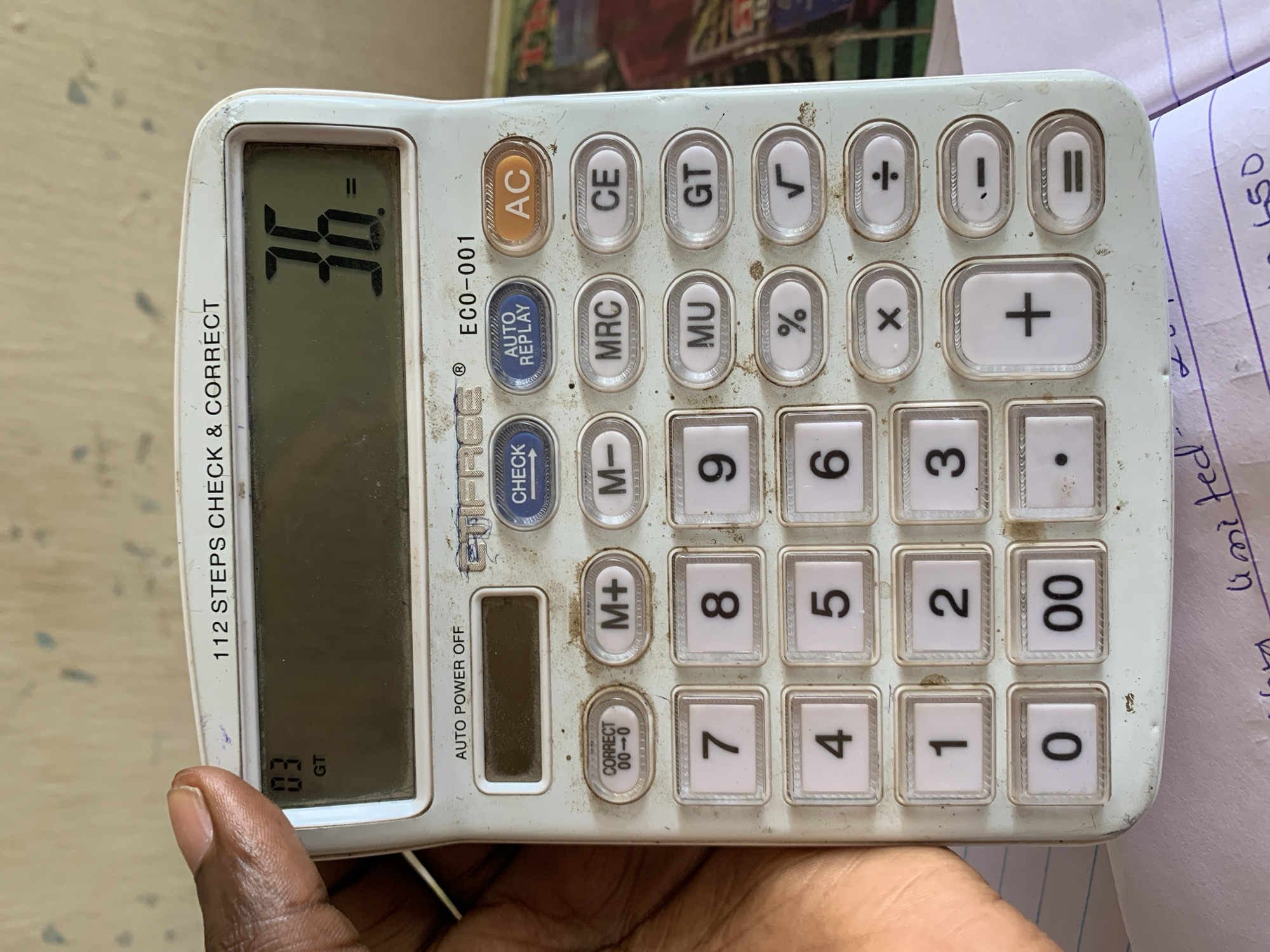 A dark-skinned hand holds a white calculator with visible wear. The calculator's display shows "36-", and "03 GT" is visible in the bottom left corner of the display. The calculator's buttons include standard functions like addition, subtraction, multiplication, division, percentage, square root, memory functions (M+, M-, MRC, MU), and numbers 0-9, along with "00". Some text on the calculator reads "112 STEPS CHECK & CORRECT," "AUTO POWER OFF," "FREE ECO-001," "CHECK AUTO REPLAY," "AC," "CE," and "GT." A piece of paper with handwritten notes is slightly visible on the right side of the calculator. There is no information to relate the image to the location of New Stadium, Nigeria.
