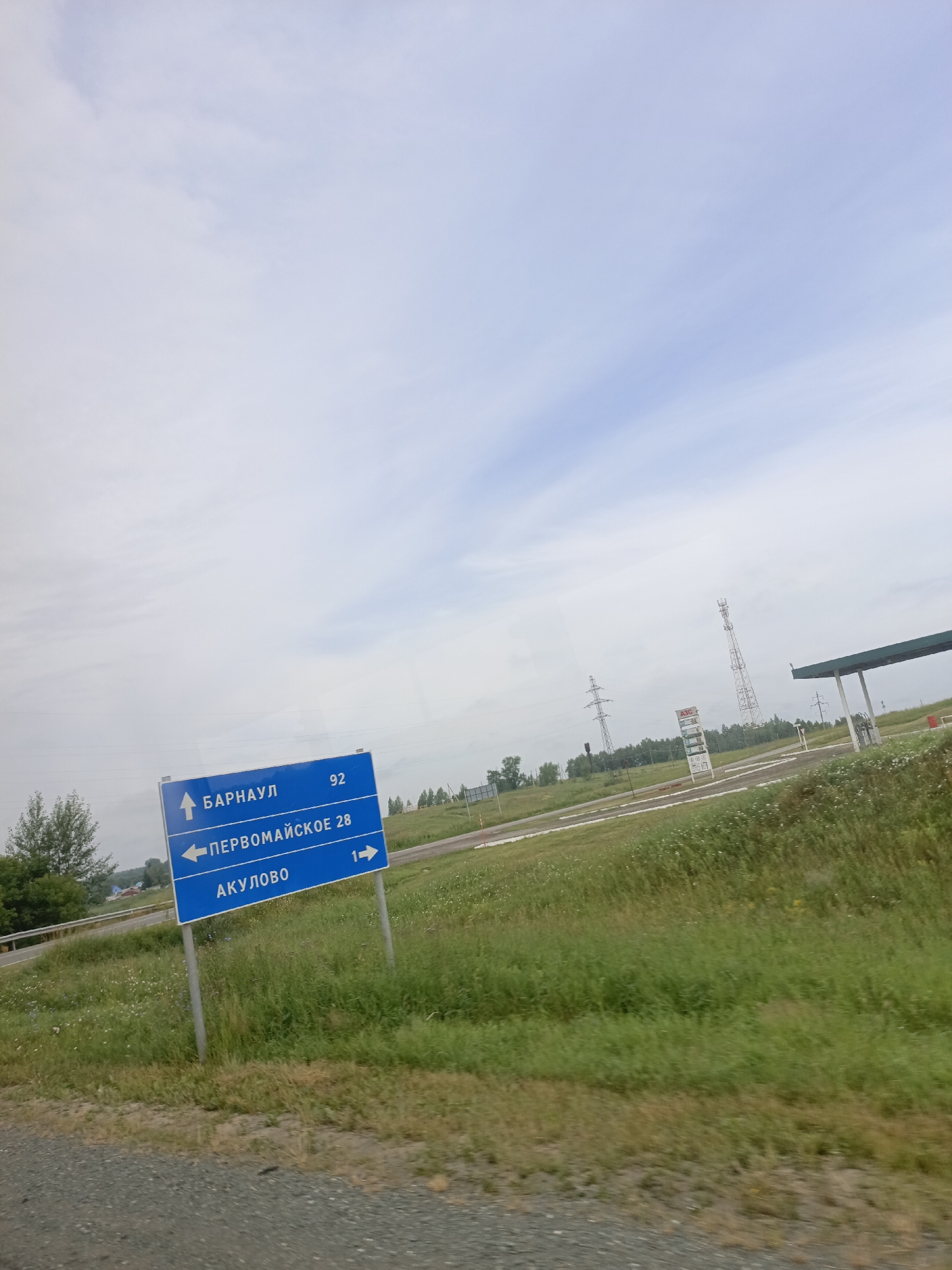 A blue road sign with white lettering is visible in the image. The sign indicates directions to "Barnaul" (92 km), "Pervomayskoye" (28 km) to the left, and "Akulovo" (1 km) to the right. The sign is supported by two metal poles. The background shows a cloudy sky, a gas station with a price board, cell towers, power lines, and a grassy field with scattered trees and wildflowers. The ground in the foreground is a gravel road. The scene appears to be a roadside location in Vidonovo, Russia.