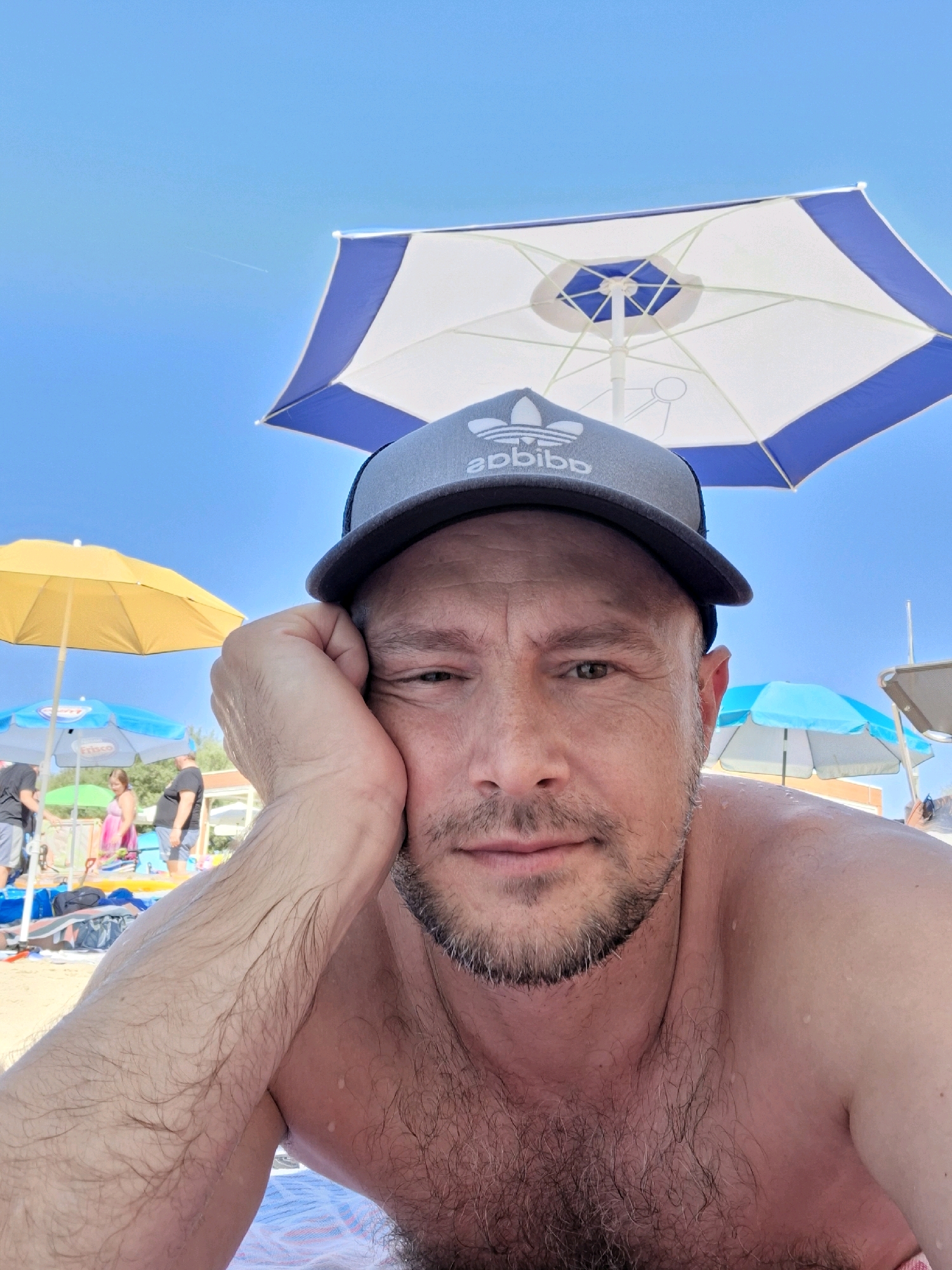 A man wearing a gray baseball cap with an Adidas logo rests his head on his hand, looking towards the camera on a sunny day at the beach in Caorle, Italy. He has a beard and is shirtless, with water droplets visible on his skin. Behind him, several beach umbrellas are visible, one yellow and two blue and white. People are scattered on the beach in the background, some standing and some sitting. The sky is clear blue.