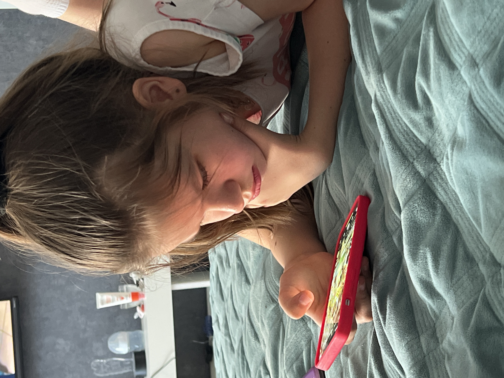 A young girl lies on a light blue textured blanket, propping her head up with her hand while holding a red-cased tablet. She's wearing a white shirt with a watermelon design. In the background, there's a white shelf with various items, including a tube of something, a bottle, and what appears to be a baby bottle. A dark gray wall is also visible. Based on the presence of what appear to be Russian household items, the location could be an apartment within Russia. However, definitively determining the specific city of Chaikovsky without further specific information or additional details is impossible.