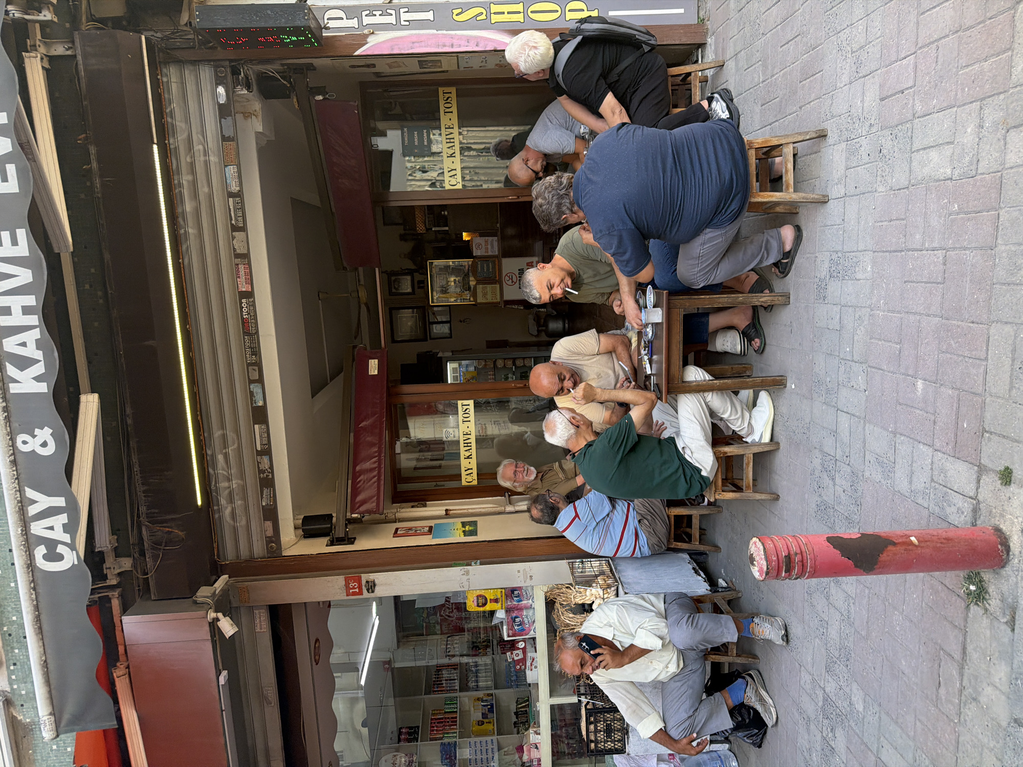 In Kadıköy, Turkey, outside a cafe with a sign that reads "ÇAY KAHVE - TOST," a group of seven men are seated at a wooden table on a sidewalk paved with brick. One man on the far left, wearing a light-colored shirt and grey pants, is speaking on his cell phone. The other six men are engaged in conversation and possibly playing a game at the table. Some are smoking. The cafe appears to be open, with shelves of goods visible through a glass storefront behind the men. A red, cylindrical bollard stands on the sidewalk in the foreground. The scene depicts a casual gathering at a local establishment on a sunny day.