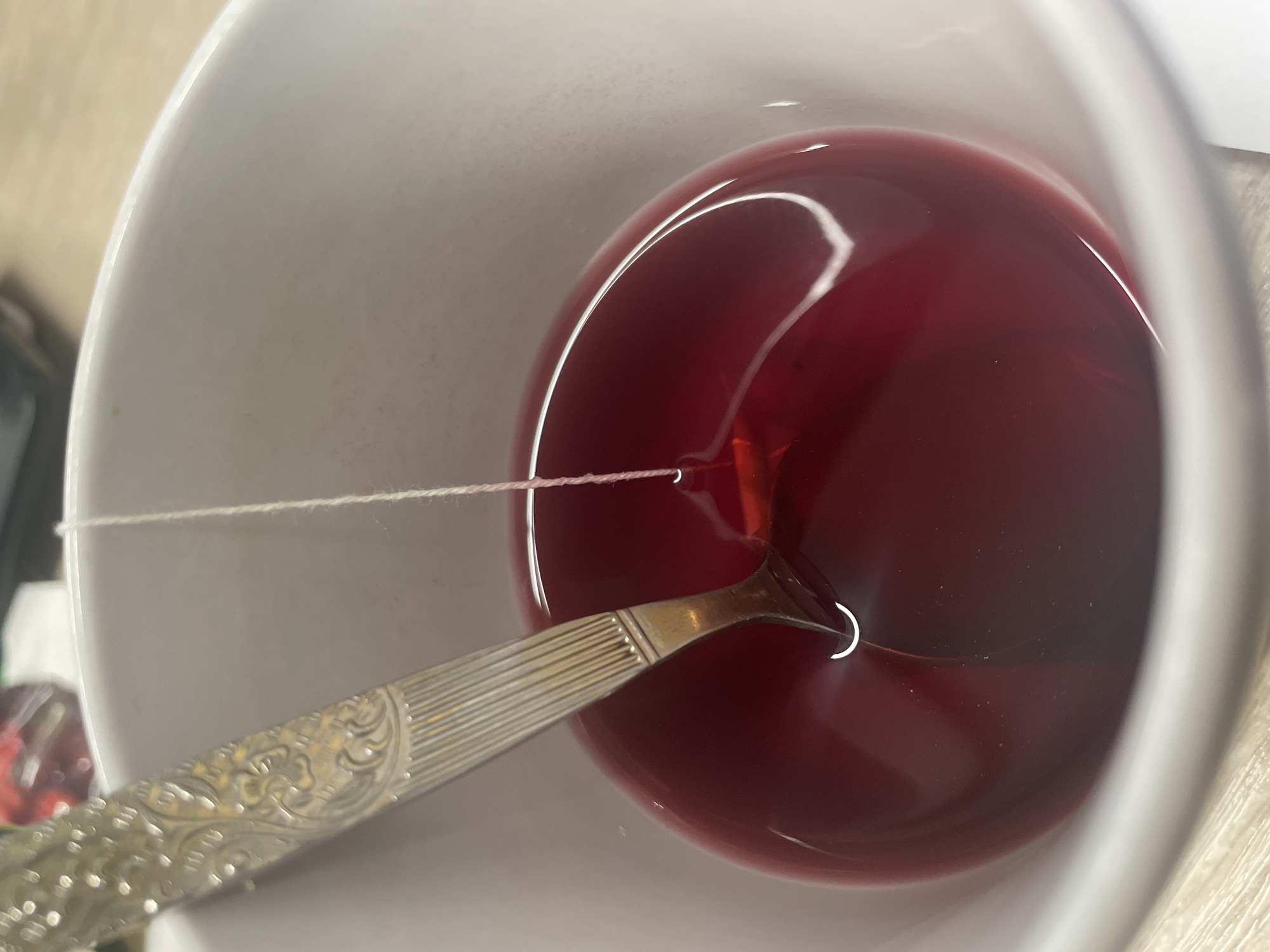 The image shows a close-up view of a white mug filled with a dark red liquid, likely tea or a beverage. A silver spoon with an ornate handle is partially submerged in the liquid, and a thin white string is suspended above the liquid, appearing to be attached to the spoon. The mug is placed on a light-colored surface, possibly a table. The background is blurred but suggests an indoor setting. There is no discernible information about the location in the city of Терволово, Russia. The image focuses on the contents of the mug and the spoon.