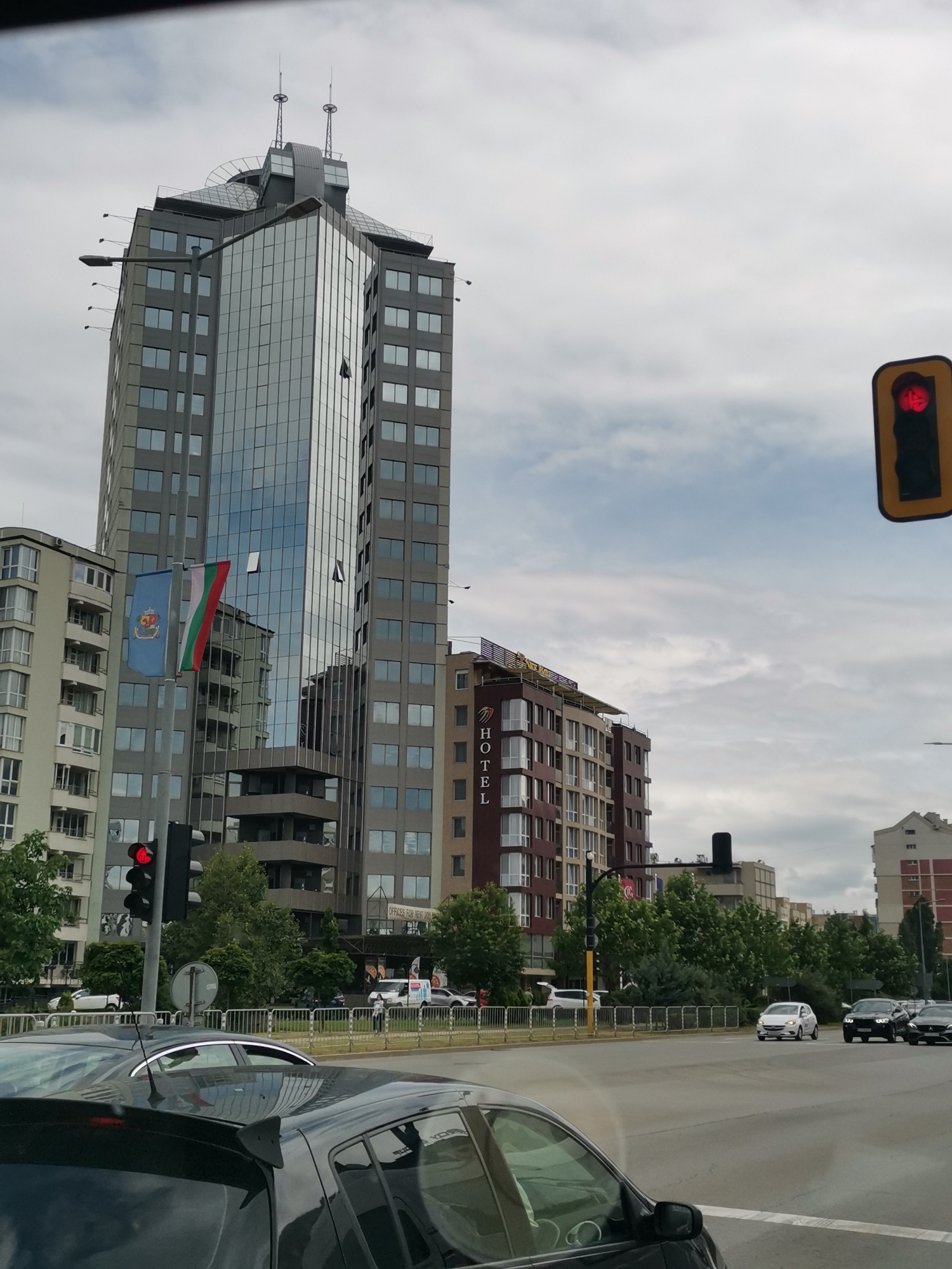 The image depicts a street view in Sofia, Bulgaria. In the background, a tall, modern skyscraper with a reflective glass facade dominates the skyline. To the left of the skyscraper, two flags are visible: one blue with a coat of arms, and the other the Bulgarian national flag. Adjacent to the skyscraper, a multi-story building displays the word "HOTEL" vertically on its facade. A traffic light with a red signal is visible on the left side of the frame. The foreground shows a street with several cars, including a dark-colored car in the immediate foreground. The sky is overcast. The scene appears to be a typical urban environment on a cloudy day.