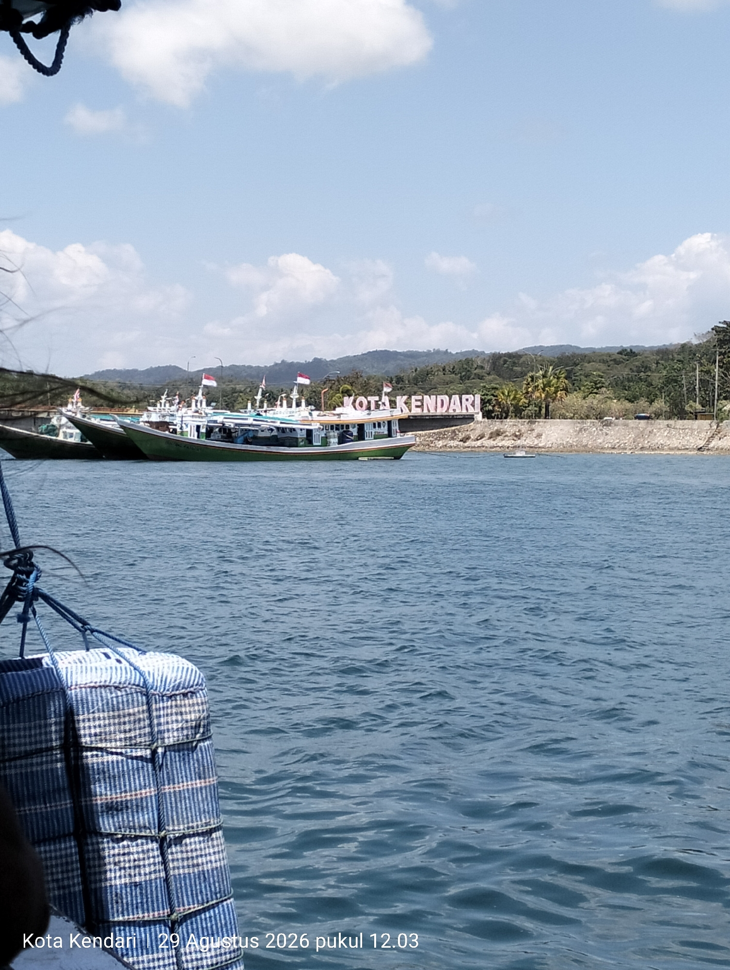 The image shows a scenic view of the Kendari Bay in Kendari City, Indonesia, on August 29, 2026, at 12:03 PM. Several fishing boats are moored in the bay, with one boat in the foreground prominently featuring a checkered pattern of blue and white fabric. In the background, a large white sign reads "KOTA KENDARI" (Kendari City), set against a backdrop of lush green hills and a clear blue sky with scattered clouds. The water is calm with gentle ripples, reflecting the bright daylight. The scene captures a typical day in this coastal Indonesian city, highlighting its maritime culture and natural beauty.