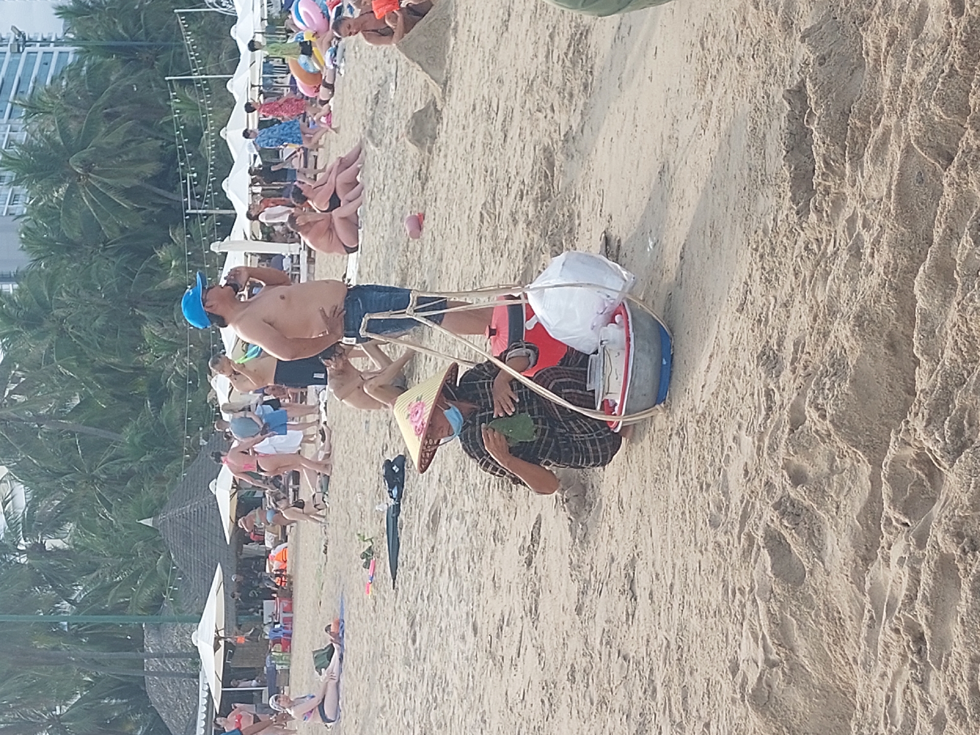 This media file depicts a beach scene in Nha Trang, Vietnam. The image is vertically oriented and the content is rotated 90 degrees clockwise. The foreground shows a person wearing a conical hat, a patterned shirt, and a face mask, crouching on the sand. They are holding what appears to be a green leaf. Beside them is a cart constructed of bamboo poles and a red and blue container, with a white plastic bag on top.
Further into the background, several other people are visible, some sitting on the sand and others walking. A row of white umbrellas and palm trees line the shore, suggesting a resort area. In the far background, a multi-story building can be seen, hinting at a more urban setting beyond the beach. The sand appears to be textured with footprints and small indentations. The lighting suggests it is daytime, possibly mid-morning or afternoon, with bright, diffused light typical of a sunny day. The overall mood is relaxed and casual, consistent with a beach environment. There is no visible text in the image.
