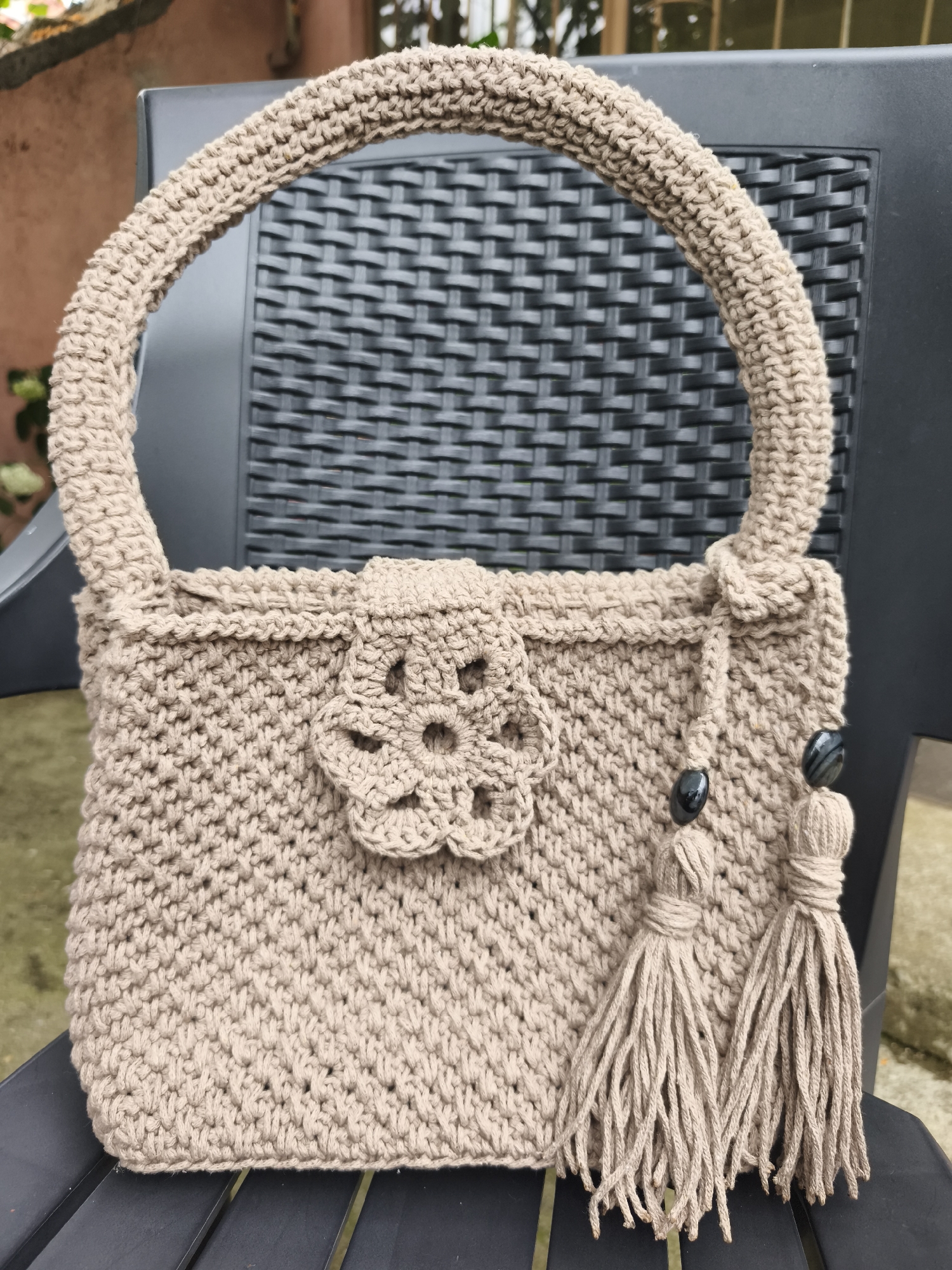 A beige, crocheted handbag is displayed on a black chair. The handbag has a rounded handle and a textured body with a floral embellishment on the front. Two tassels with dark, striped beads hang from the front of the bag. The background shows a hint of an outdoor setting with a wall and greenery. The image appears to be taken in daylight. The location context is Marinka, Bulgaria.