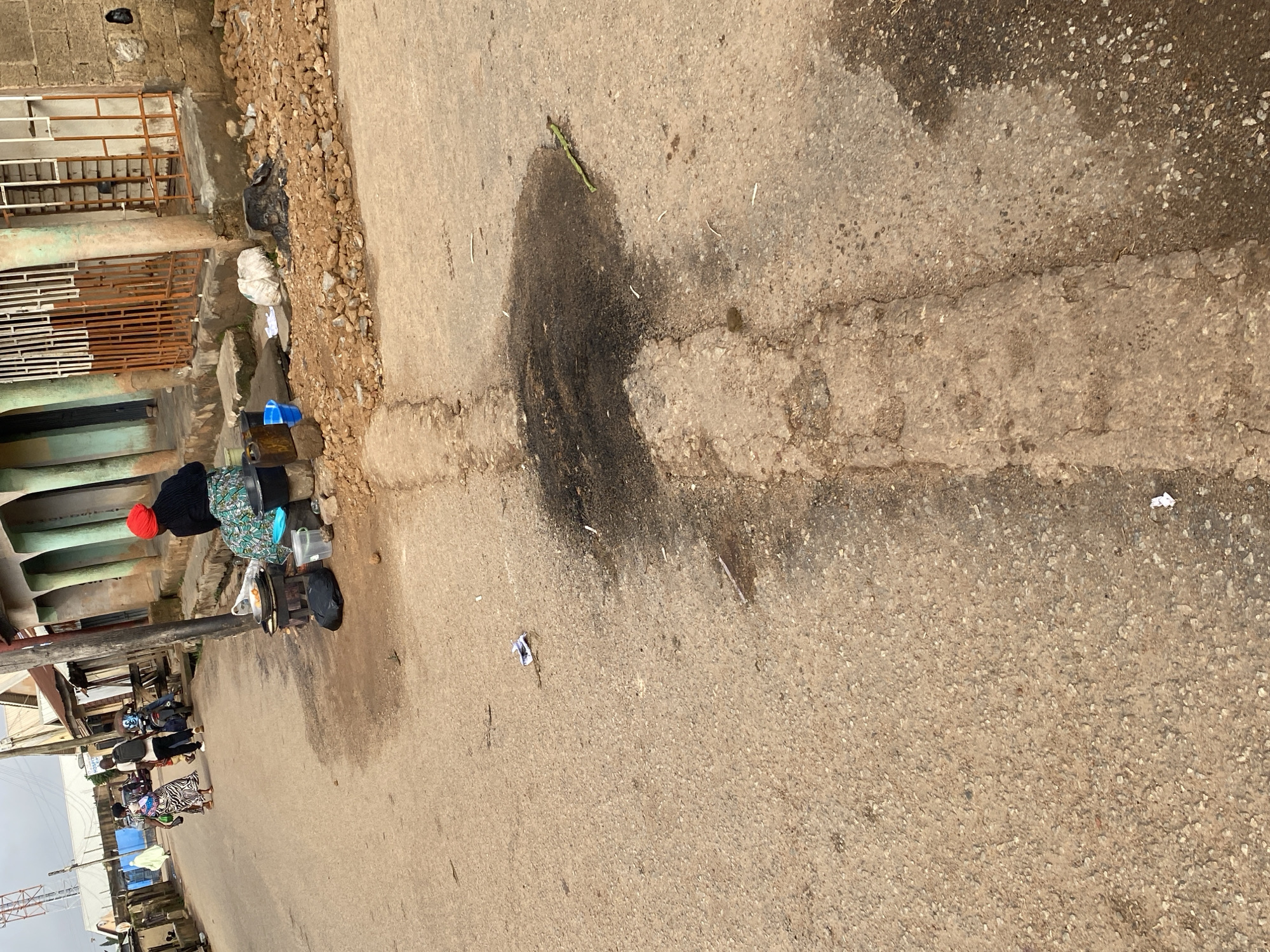 The image depicts a street scene in Jos, Nigeria. The foreground is dominated by a cracked asphalt road with a dark, oily stain. A narrow, raised concrete channel runs lengthwise through the road. In the middle ground, a woman wearing a red headscarf and a patterned skirt sits on a low wall next to a small stove with a pot on it. Various items, including plastic containers and a black garbage bag, are placed around her. Behind her, a few people are walking down the street, and a motorcycle is parked. The background shows buildings with corrugated metal roofs and a tall communication tower. The environment appears to be an urban or semi-urban setting with some signs of disrepair and everyday commerce.
