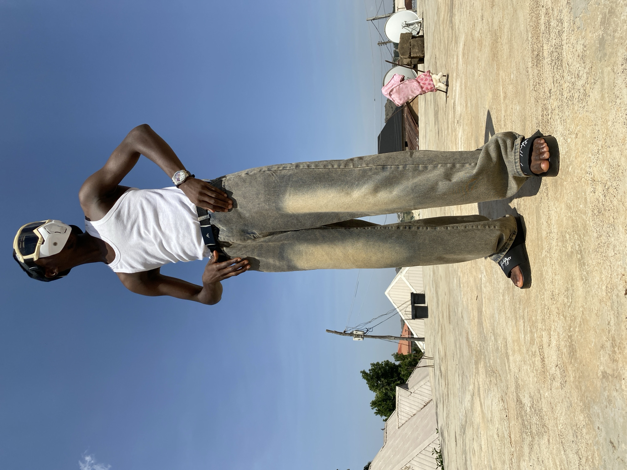A young Black man is standing on a concrete rooftop under a bright blue sky in Bukuru, Nigeria. He is wearing a white tank top, greyish-brown jeans, black sandals, a watch, and a white face mask with dark accents. His hands are placed near his hips. In the background, there are satellite dishes, rooftops, a pink child's dress hanging over the roof's edge, utility poles, trees, and other buildings. The location appears to be a residential area. The image is rotated 90 degrees counter-clockwise.