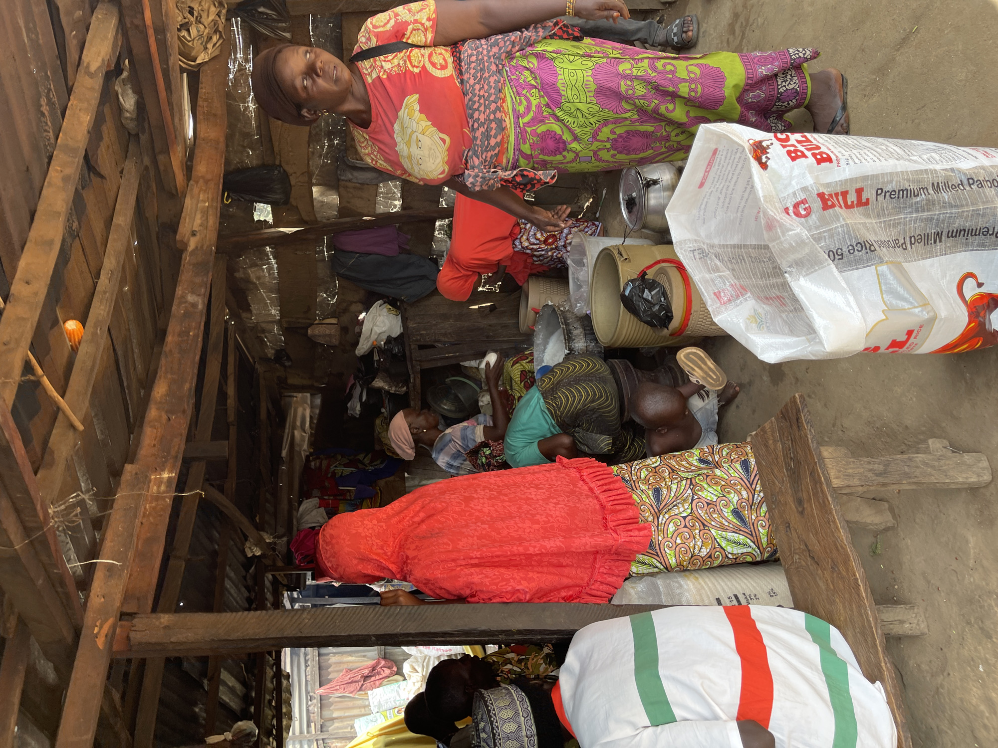 This is a medium shot, taken at eye-level, of an open-air market stall in Abuja, Nigeria. The stall has a corrugated metal roof supported by rough-hewn wooden beams. The floor is packed dirt. Several people are present. In the foreground on the right, a woman with dark skin and a brown headscarf faces away from the camera, looking towards the left. She wears a bright red t-shirt with a gold graphic and a long, brightly colored wrap skirt with green and pink floral patterns. Her right arm is bent, and her hand is near her waist. In front of her is a large, translucent plastic bag with red lettering that reads "BIG BULL Premium Milled Pâté Rice 50". To her left, a woman in a vibrant red headscarf and patterned skirt stands facing away from the camera. To her left and further back, a woman in a green top and patterned skirt is crouched, attending to something on the ground. Several large buckets and containers are visible within the stall. In the far left of the image, a person wearing a white shirt with red and green stripes stands facing left. Other individuals are partially visible in the background, suggesting a bustling market environment.