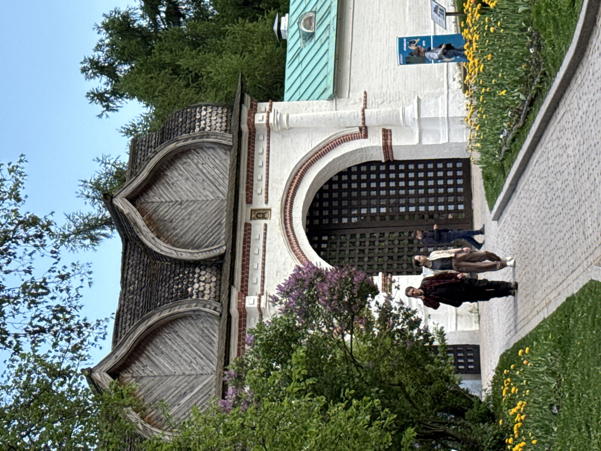 The image depicts the Front Gate of the Kolomenskoye Estate in Moscow, Russia. The gate is a white stone structure with a large, arched wooden doorway. The roof is made of wooden shingles arranged in curved patterns. In front of the gate, a group of three people walk along a stone pathway. To the right of the pathway is a grassy area with yellow flowers. A lilac bush is in the foreground, partially obscuring the base of the gate. The background features trees and a glimpse of the sky.