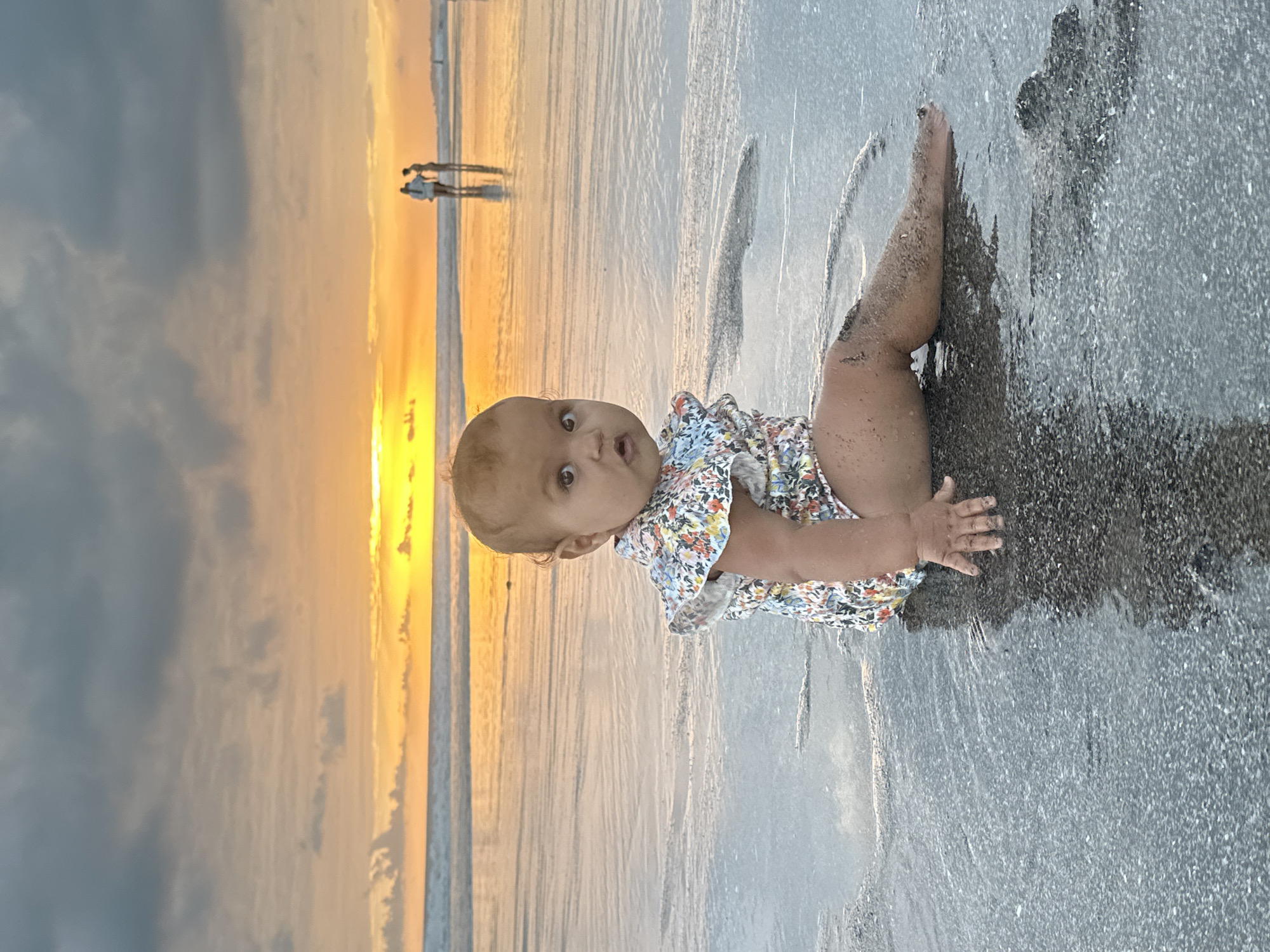 A young child with a floral swimsuit sits on a wet, dark sand beach in North Kuta, Indonesia, during sunset. The child's legs are covered in sand and partially submerged in shallow water. The child is looking towards the camera with wide eyes. In the background, the ocean reflects the vibrant orange and yellow hues of the setting sun, with a few silhouetted figures walking along the shoreline. The sky is a mix of dark grey clouds and bright sunset colors.