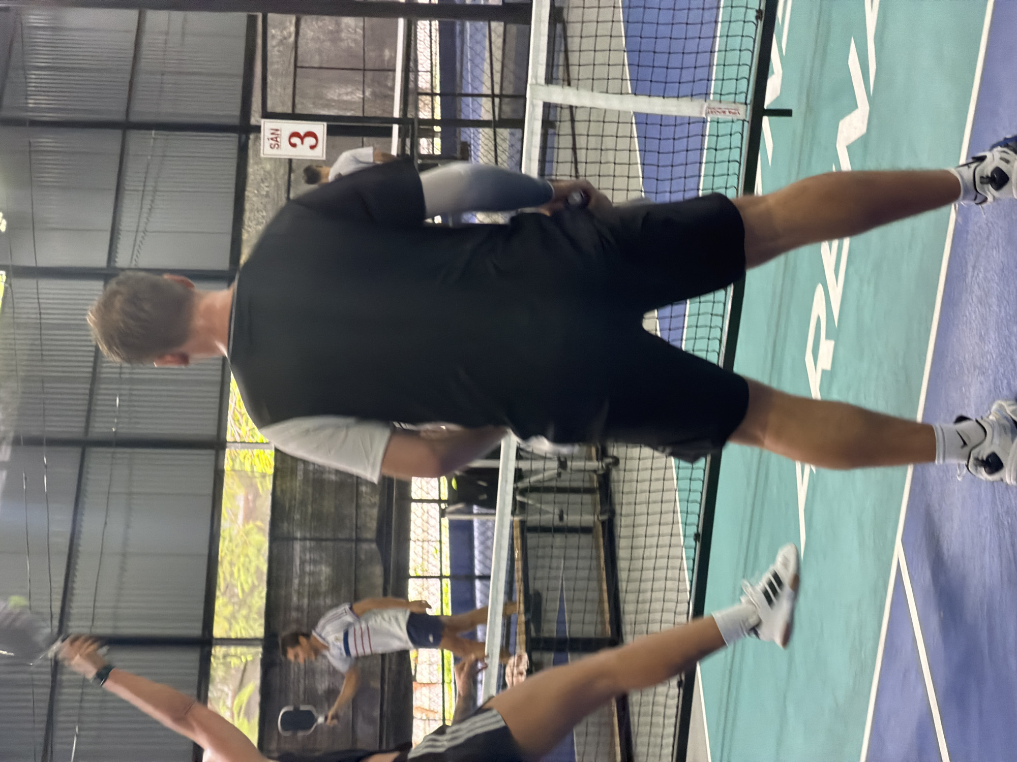 The image depicts an active pickleball game taking place at an indoor or semi-enclosed sports facility in Nam Nha Trang Ward, Vietnam.
Several individuals are visible on the court, engaged in play. In the immediate foreground, a person is seen from behind, wearing a black t-shirt, black shorts, and an arm sleeve, holding a pickleball paddle. Their white and black athletic shoes are prominent on the purple section of the court. Another player's arm and paddle are visible to the left, indicating more participants in the foreground.
The court itself is brightly colored with distinct teal and purple sections, marked by white lines, and divided by a white net. In the background, another player, wearing a white and blue striped shirt and white shorts, is also holding a paddle, suggesting active play on the far side of the net.
The setting features corrugated metal walls, indicative of an industrial or purpose-built sports structure. Natural light filters in from outside, where green foliage is visible, suggesting either an open-air facility or one with large windows or openings.
A notable detail is a white sign with a red border, clearly displaying the text "SÂN 3". "SÂN" translates to "Court" or "Field" in Vietnamese, indicating this is Court number 3. The scene captures the dynamic atmosphere of a pickleball match, likely during daytime hours.