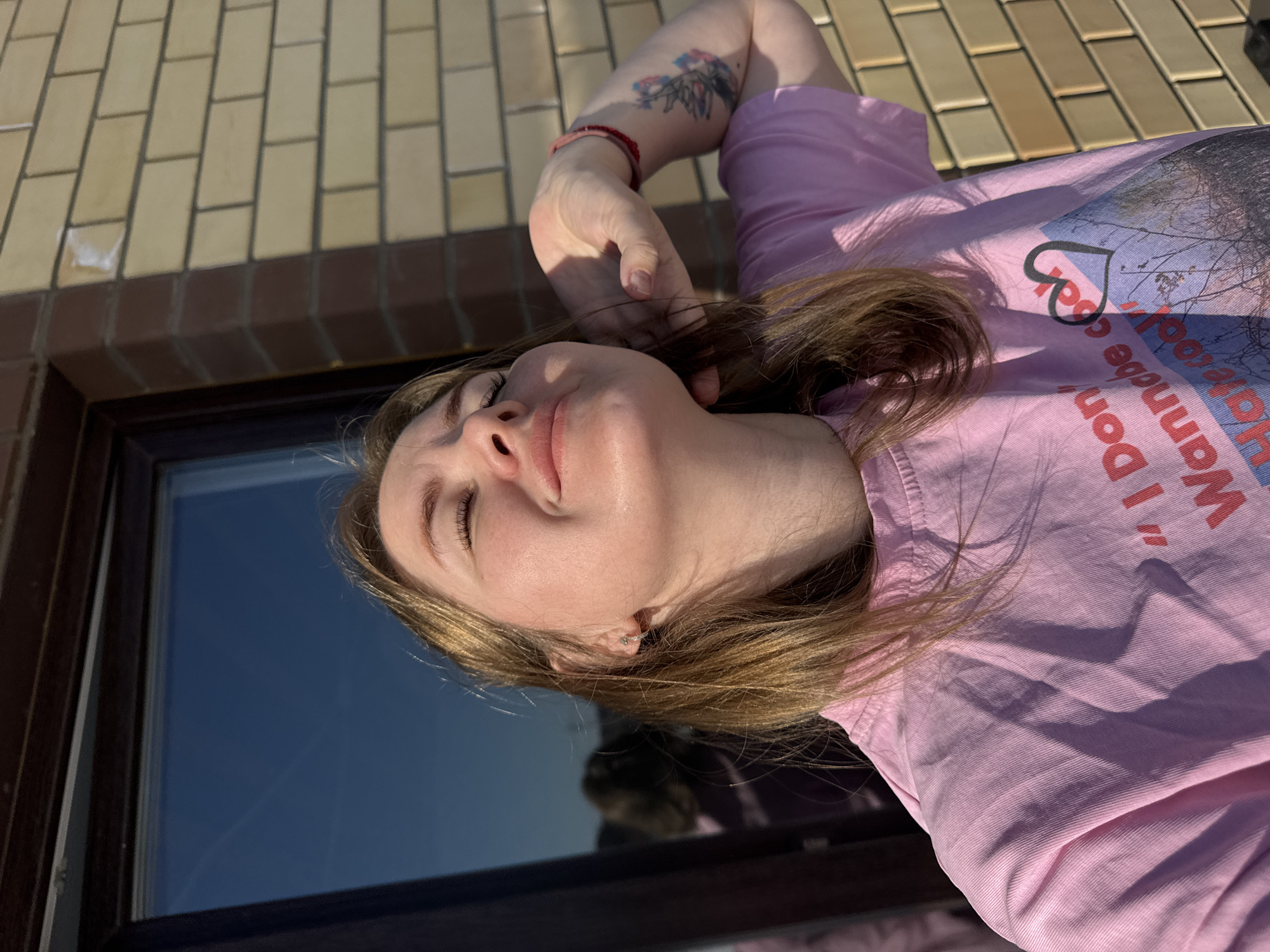 The image depicts a female subject from the chest up, positioned outdoors in Gornyi (Dvurechenskoe S/P), Russia. The subject has light brown hair, eyes closed, and her head tilted backward with a slight smile, appearing to face upwards towards sunlight. She wears a pink short-sleeved t-shirt with the text "I Don't Wannabe cool" and a graphic featuring a black heart above a landscape. A silver stud earring is visible in her left ear. Her right arm is bent with the hand positioned behind her neck, revealing a red beaded bracelet on the wrist and a multi-colored floral tattoo on the upper arm. The background consists of a light yellowish-brown brick wall on the right and a dark-framed window on the left, reflecting a clear blue sky and a partial, blurred silhouette of the subject. Strong direct sunlight is evident, casting prominent shadows.