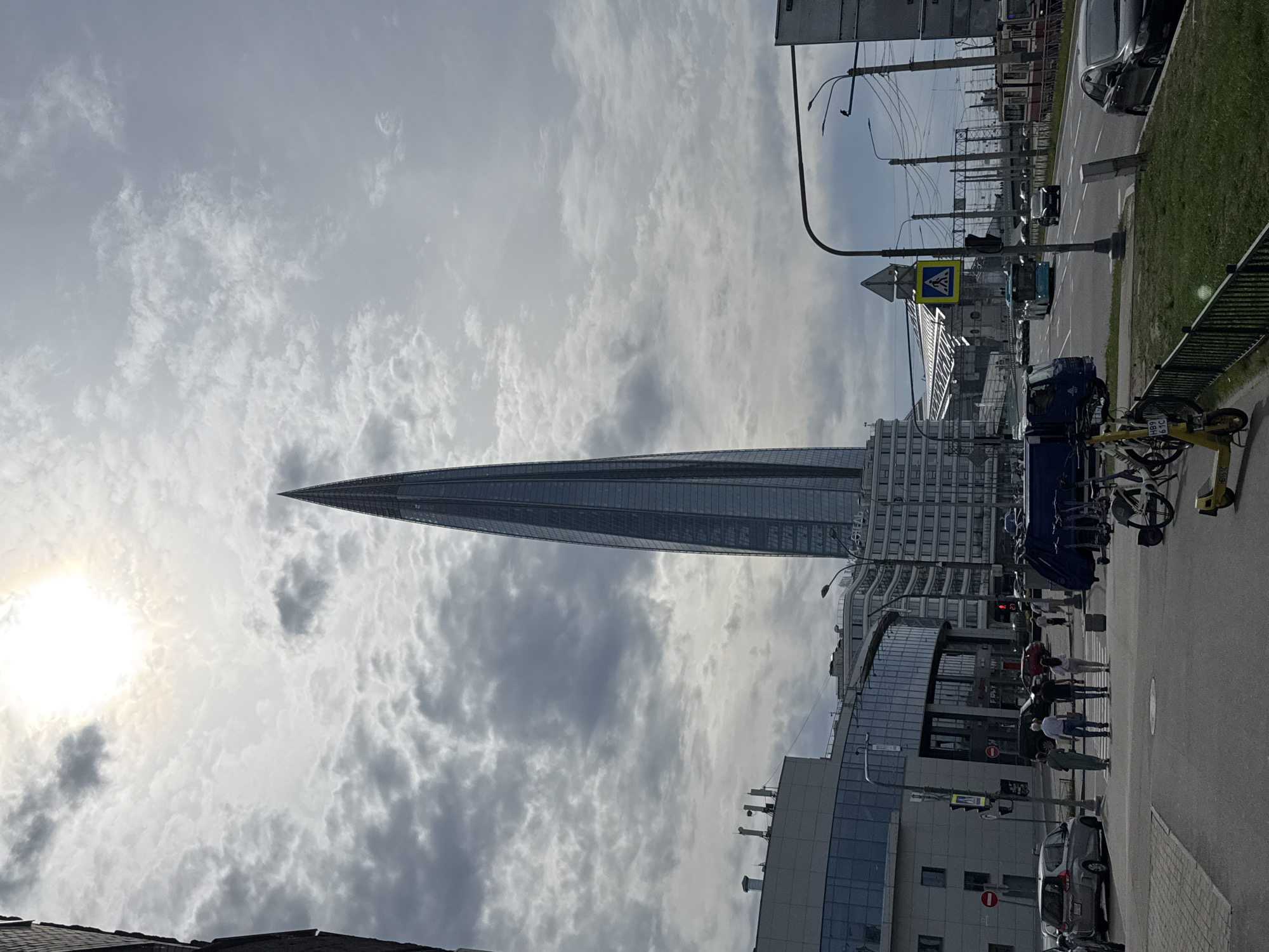 The image shows the Lakhta Center skyscraper in Saint Petersburg, Russia, on a cloudy day. The building, a prominent landmark, has a distinctive spire shape and a glass facade. The sun is visible through the clouds, creating a diffused light. Below the skyscraper are several other buildings, a street with pedestrian crossings, traffic lights, and parked cars. Some pedestrians are crossing the street at a designated zebra crossing. Bicycles and scooters are parked on the sidewalk. A street light and overhead electrical wires are visible above the street.