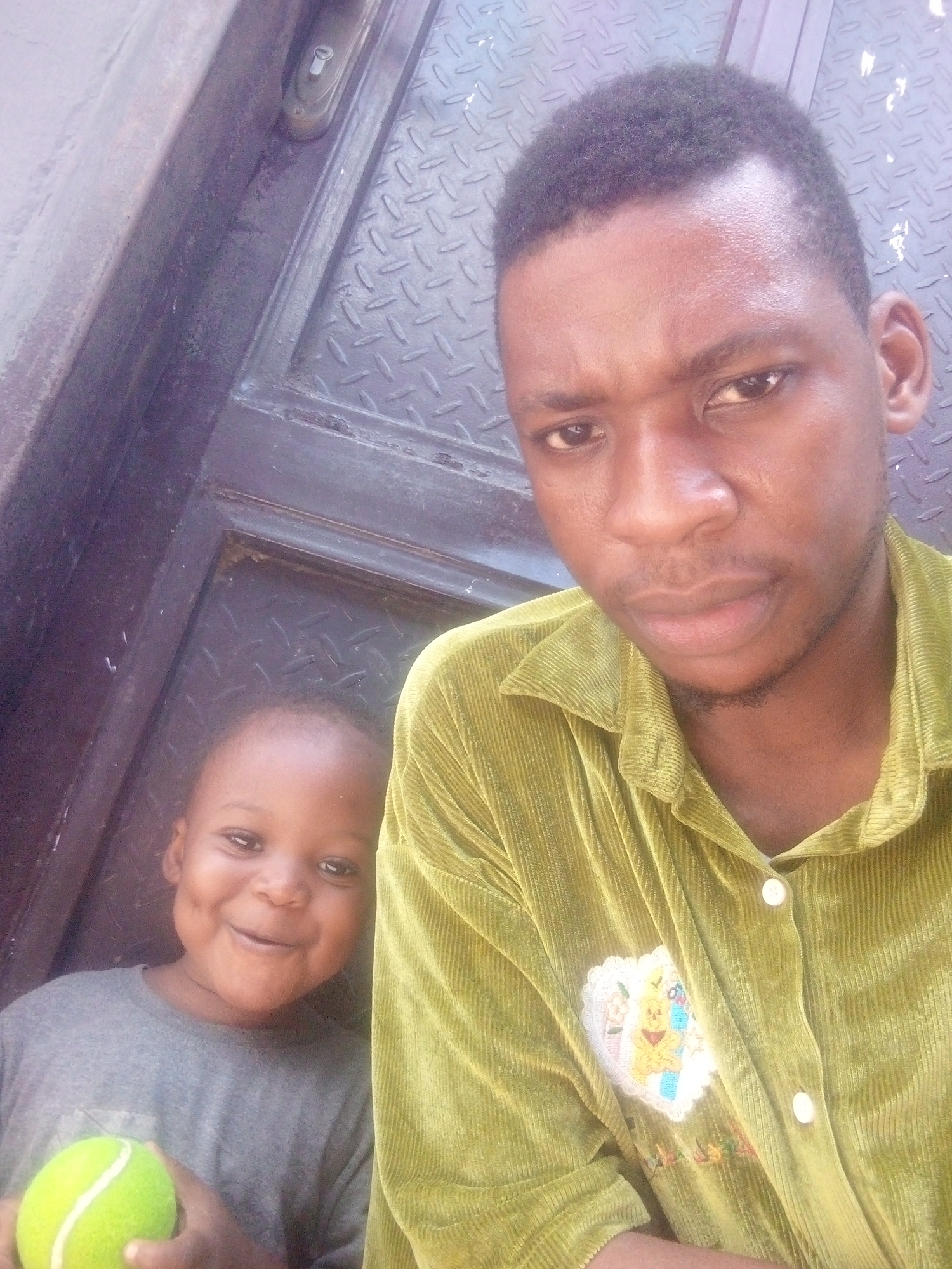 An adult male and a young child are captured in a close-up, slightly tilted image, appearing to be a selfie. The adult, with dark skin, short dark hair, and subtle facial hair, occupies the right side and center of the frame. He wears a light green, possibly corduroy or velvet, long-sleeved collared shirt with white buttons. An embroidered patch, featuring a light-colored, possibly cartoon character (resembling Winnie the Pooh) within a heart-shaped border, is visible on his left chest area. His expression is neutral to slightly serious.
To the left and slightly behind the adult, a young child with dark skin is partially visible. The child is smiling widely, revealing dimples, and appears joyful. They are wearing a grey long-sleeved shirt and are holding a bright neon green tennis ball with white lines.
The background consists of a dark, textured surface, likely a metal door or wall with a diamond plate (also known as checker plate) pattern. A dark frame or adjacent wall section is visible to the far left. A small, metallic lock or latch mechanism is discernible at the top of the textured surface. The lighting appears bright, suggesting it is daytime. No discernible text is visible on any objects or surfaces.