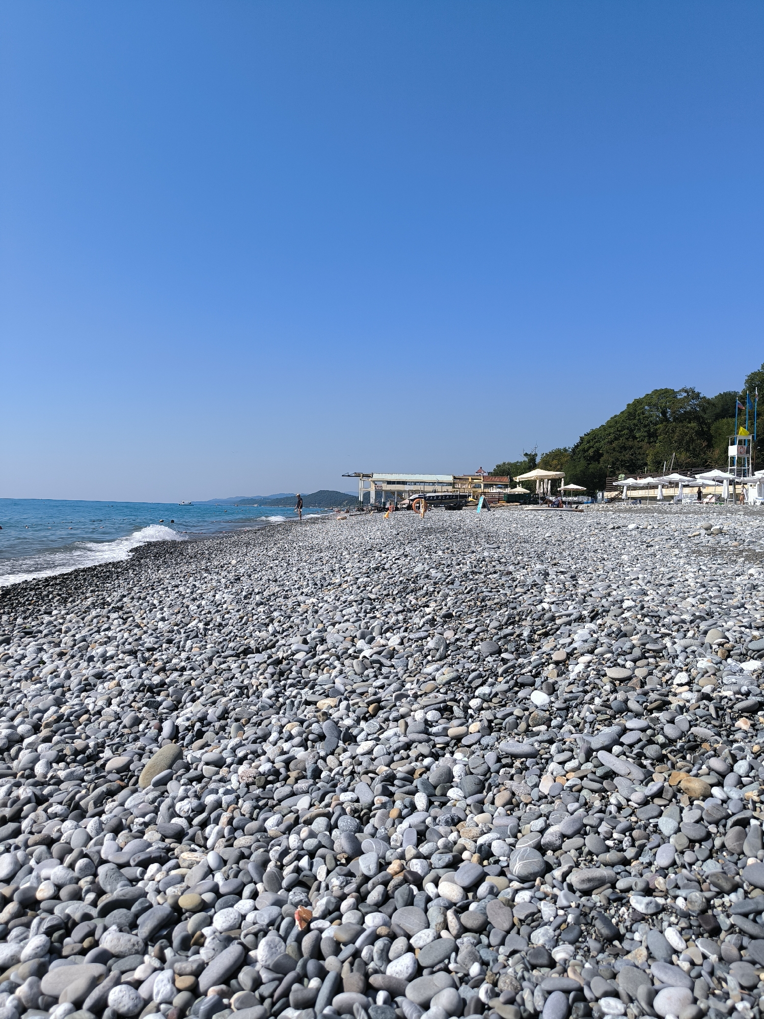 This is a wide-angle, eye-level, outdoor, daytime photograph of a pebble beach in Sochi, Russia, under a clear blue sky. The foreground is dominated by a dense, textured expanse of smooth, gray pebbles of varying sizes. In the middle ground, the gentle waves of the Black Sea lap at the shore, creating a white foam line. Several people are visible scattered across the beach, some walking and others appearing to be relaxing under white beach umbrellas.
Further back, a cluster of buildings lines the coast, including a long, low-rise structure with a covered seating area, and a multi-story building that appears to be a cafe or restaurant. White tents and lounge chairs are set up on the sand. Behind the buildings, a lush green hillside covered in trees rises steeply. A tall lifeguard tower with flags is visible on the right side of the frame. The horizon line is low, with distant mountains visible in the haze. A white boat is visible on the water in the distance. The overall impression is one of a peaceful summer day at a popular seaside resort.