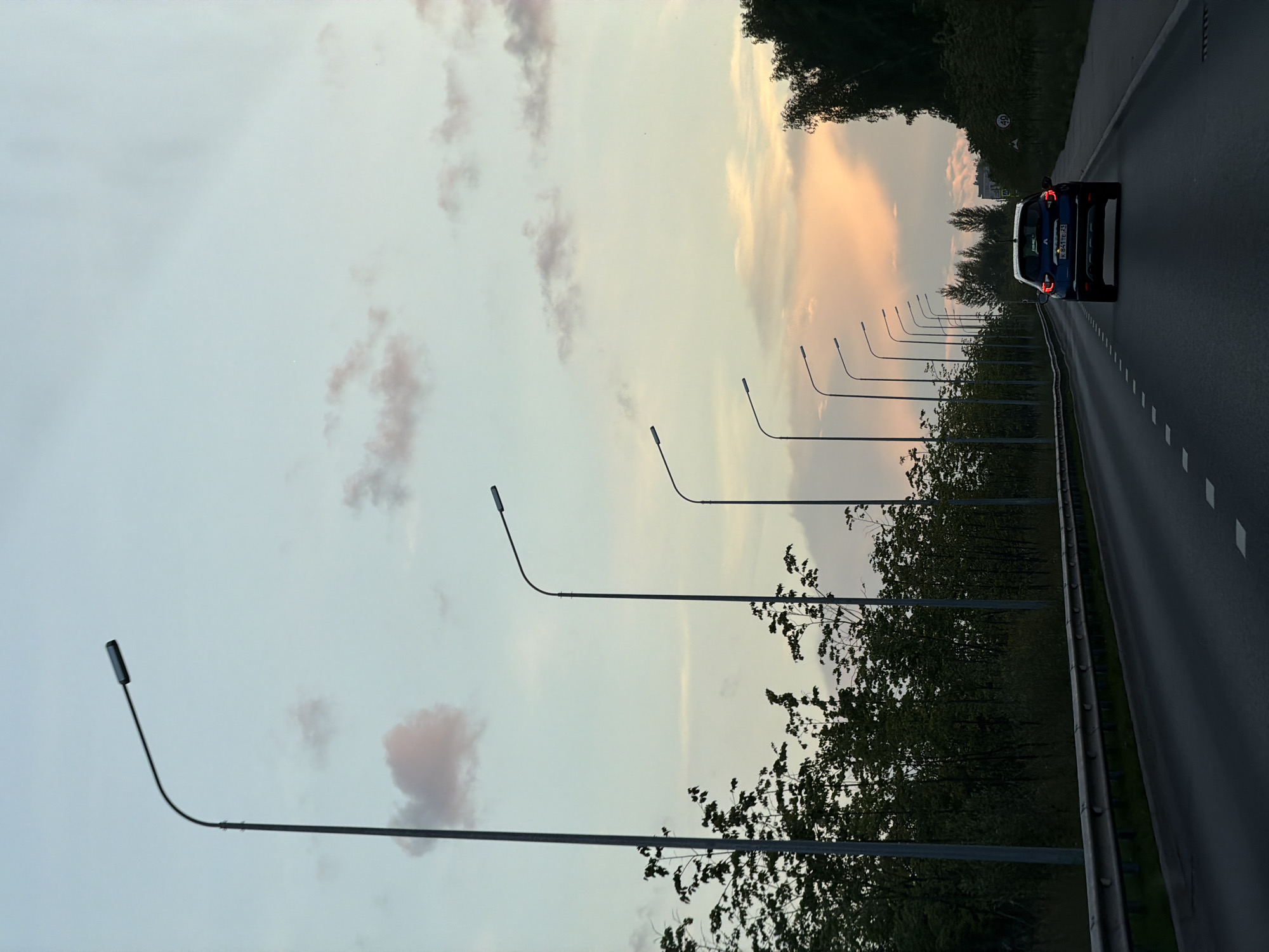The image captures a street scene at dusk in Kazan, Russia. A dark blue Renault Captur SUV is traveling away from the camera on a multi-lane road. The sky is a soft gradient of pale blue and pink, with scattered clouds. A row of streetlights lines the left side of the road, with trees and dense foliage behind them. A metal guardrail runs along the edge of the road. The road surface is asphalt with white lane markings. On the right side of the road, there is a sidewalk, more trees, and a speed limit sign indicating 40. In the distance, there are some buildings visible, suggesting an urban or suburban environment.