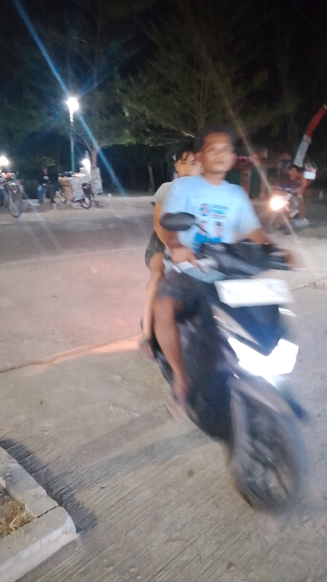 The image depicts a nighttime scene in Karanglangu, Indonesia, likely on a street or pathway. In the foreground, two individuals are riding a dark-colored motorcycle. The person in the front appears to be an adult, wearing a light blue t-shirt with a graphic design. They are operating the motorcycle. Behind them, a child, likely a girl, is seated. She has dark hair and is looking towards the front. Both individuals are somewhat blurred due to motion. The motorcycle has its headlight on, illuminating the paved surface ahead. In the background, trees are visible against a dark sky, and a streetlamp with a bright light source is present. Other indistinct figures and parked motorcycles are also visible in the middle ground. The ground is paved and appears somewhat weathered.