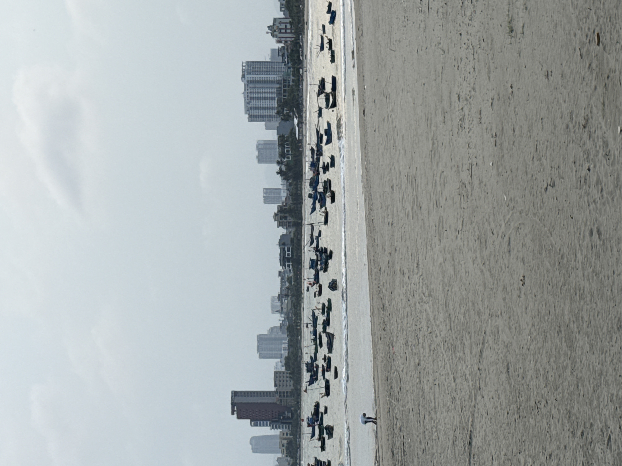 The image shows a view of Mỹ Khê Beach in Đà Nẵng, Vietnam. The beach is lined with umbrellas and chairs. A person is standing on the beach, possibly near the water's edge. In the background, there are high-rise buildings, likely hotels and apartments. The sky is overcast, with visible clouds. The beach is mostly flat and sandy.