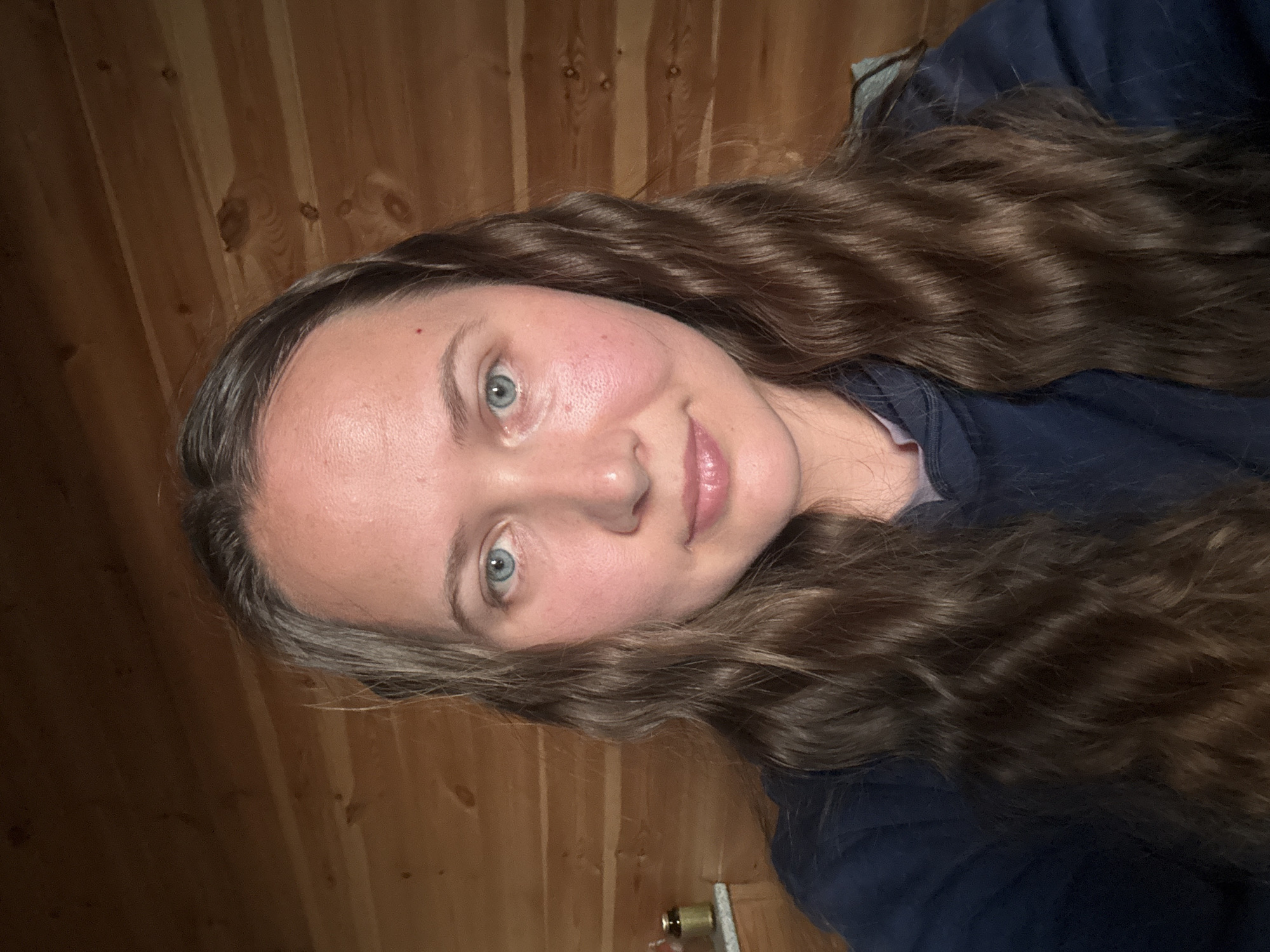 A woman with long, wavy brown hair and blue eyes is facing the camera. She is wearing a dark blue top. The background consists of wooden planks, likely a wall. The setting is not explicitly identified, and there is no indication that this photo was taken in Tyulyuk, Russia.