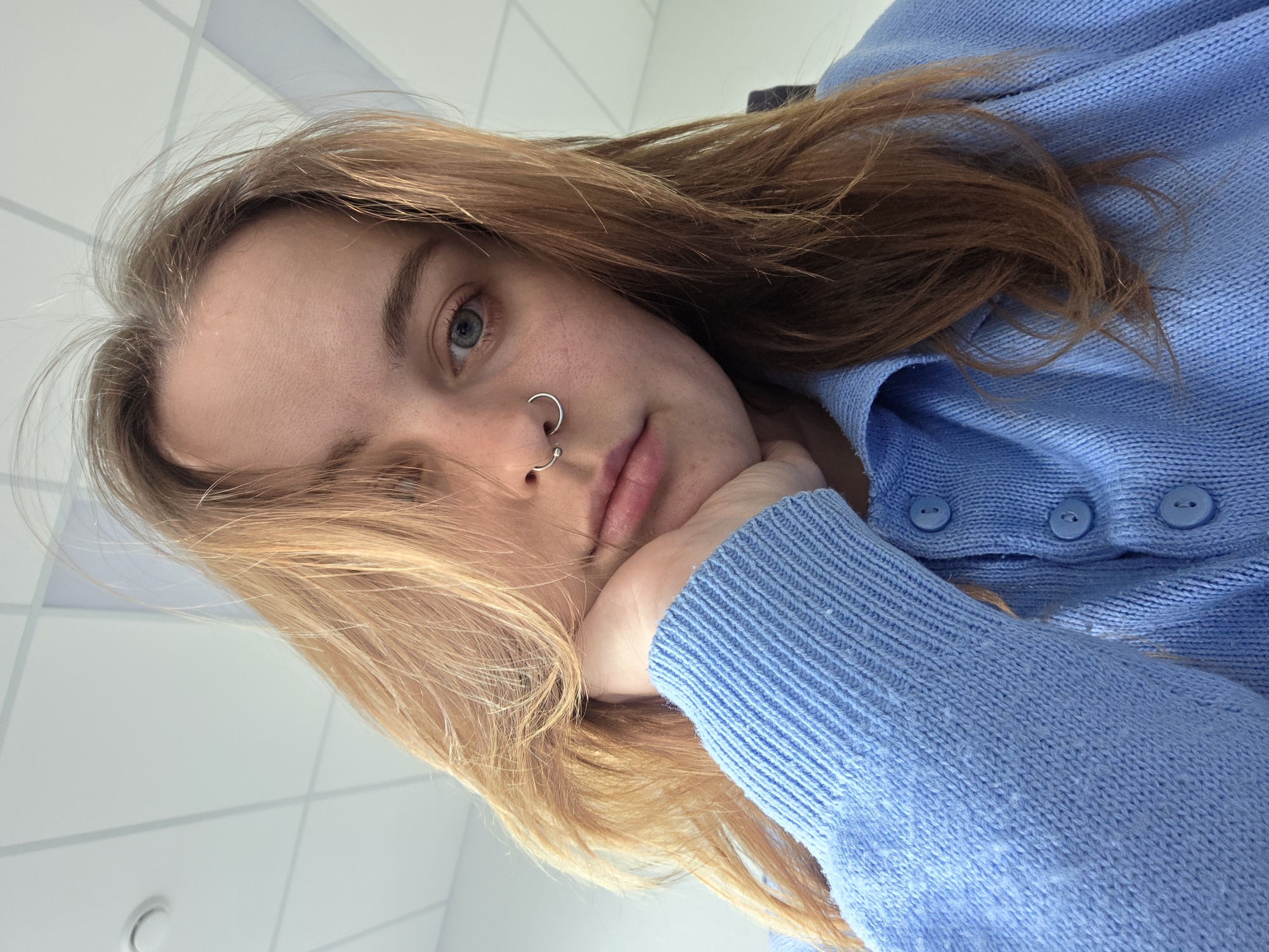 A close-up, low-angle shot depicts a person with fair skin, light brown hair, and blue eyes. The individual has a silver nose ring in their left nostril. They are wearing a light blue knit sweater with buttons. Their head is resting on their left hand. The background consists of a white ceiling with a grid pattern. The location is Unknown, Unknown.