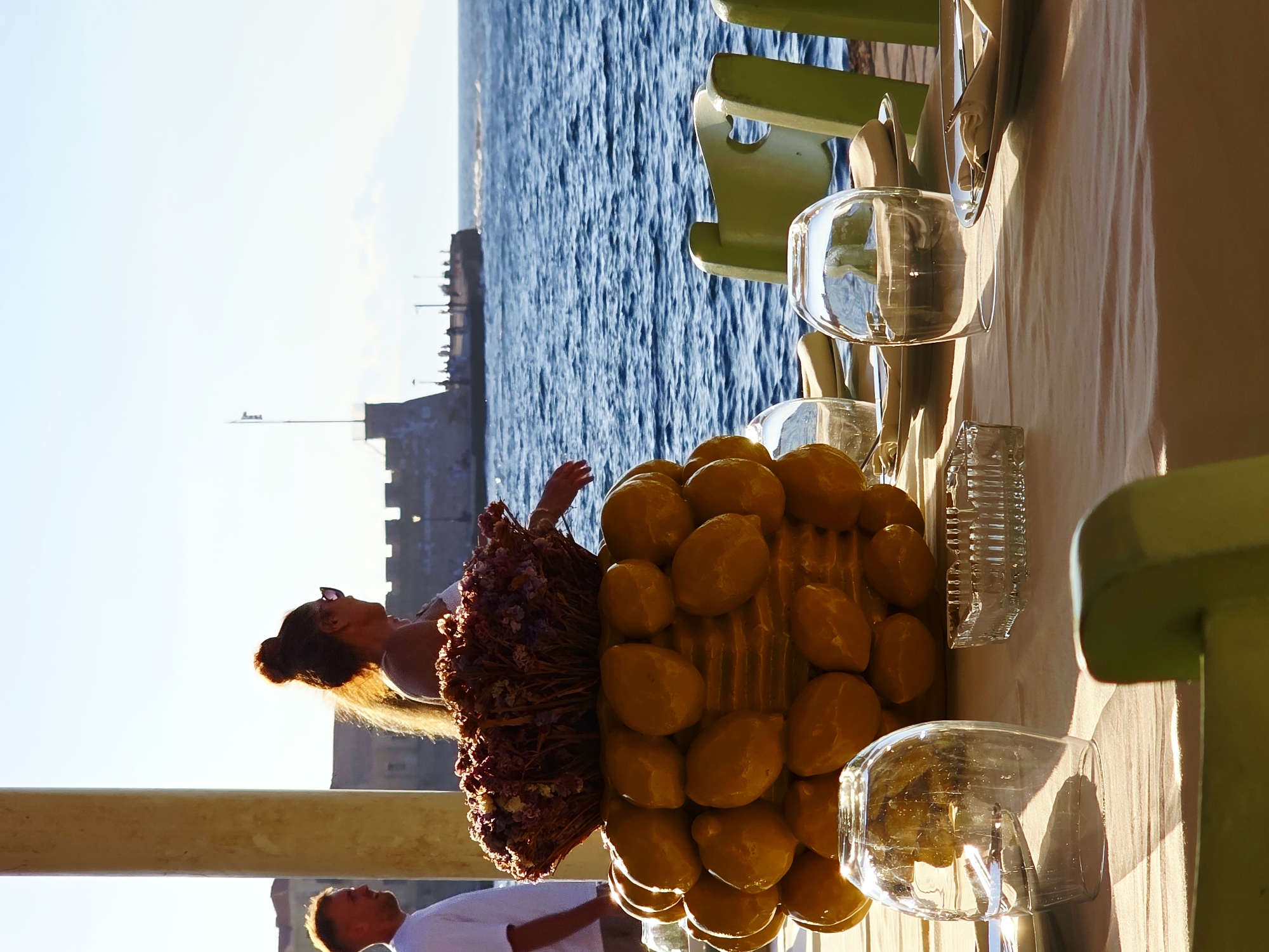 In Chania, Greece, a table is set for dining outdoors overlooking the sea. A centerpiece featuring a basket of dried flowers sits atop a large, decorative arrangement of faux yellow lemons. In the foreground, a tablecloth covers the table, with clear glasses, plates with folded napkins, and an ashtray visible. To the left, a man in a white shirt stands near a pillar, while to the right, a woman with her hair in a bun, wearing sunglasses, is looking out towards the water. The background shows the blue sea, a stone pier with people, and a distant structure. The lighting suggests it is a sunny day.
