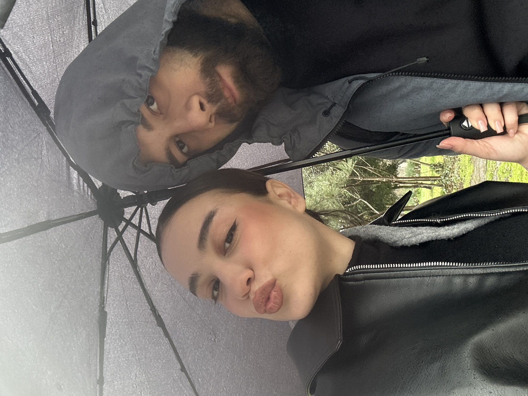 A selfie-style image shows two people under an umbrella. The person in the foreground has brown hair pulled back, light skin, and is wearing a black leather jacket with a zippered closure. They are making a kissing face towards the camera. Behind them, a person with a beard and a gray hooded jacket is looking at the camera. The background is blurry but appears to show green trees. The location cannot be determined as Sofia based on the image's visual content.