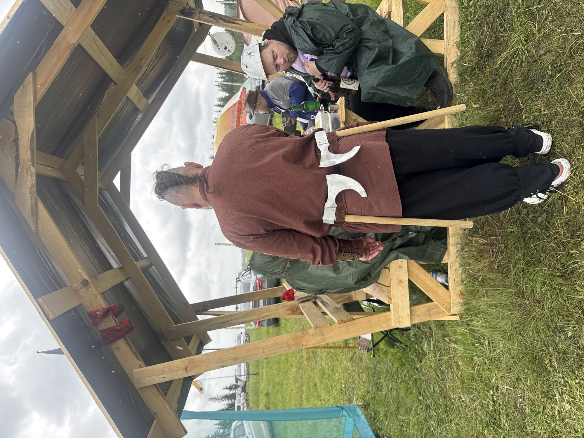 This image depicts an outdoor gathering, likely a festival or historical reenactment, in Большая Кивара, Russia, under an overcast sky.
The scene is set around a simple, unpainted wooden structure, possibly a temporary shelter or stall, which has a black tarp serving as its roof. Bits of red fabric or socks are hanging from one of the wooden beams inside the structure.
In the foreground, with his back largely to the viewer, is a man wearing a brown tunic and black baggy pants, paired surprisingly with modern white sneakers featuring black laces. Two large, ornate axes are tucked into a belt or sash at his lower back, their metal heads prominently displayed. He has short hair with a small braid or topknot.
Behind him, another man looks directly at the camera with a casual, almost amused expression. He is dressed in a dark green raincoat over a light-colored hoodie and wears a white hard hat. He holds a brown glass bottle, possibly containing an alcoholic beverage. A third person is partially visible behind him, also holding a bottle.
The ground is a grassy field, suggesting a rural or semi-rural outdoor setting. In the mid-ground and background, other tents, parked cars, and additional simple wooden constructions are visible, reinforcing the impression of a large outdoor event or camp. The sky is bright but entirely overcast, indicating daytime and potentially damp weather, consistent with the man's raincoat. No discernible text is visible in the image.