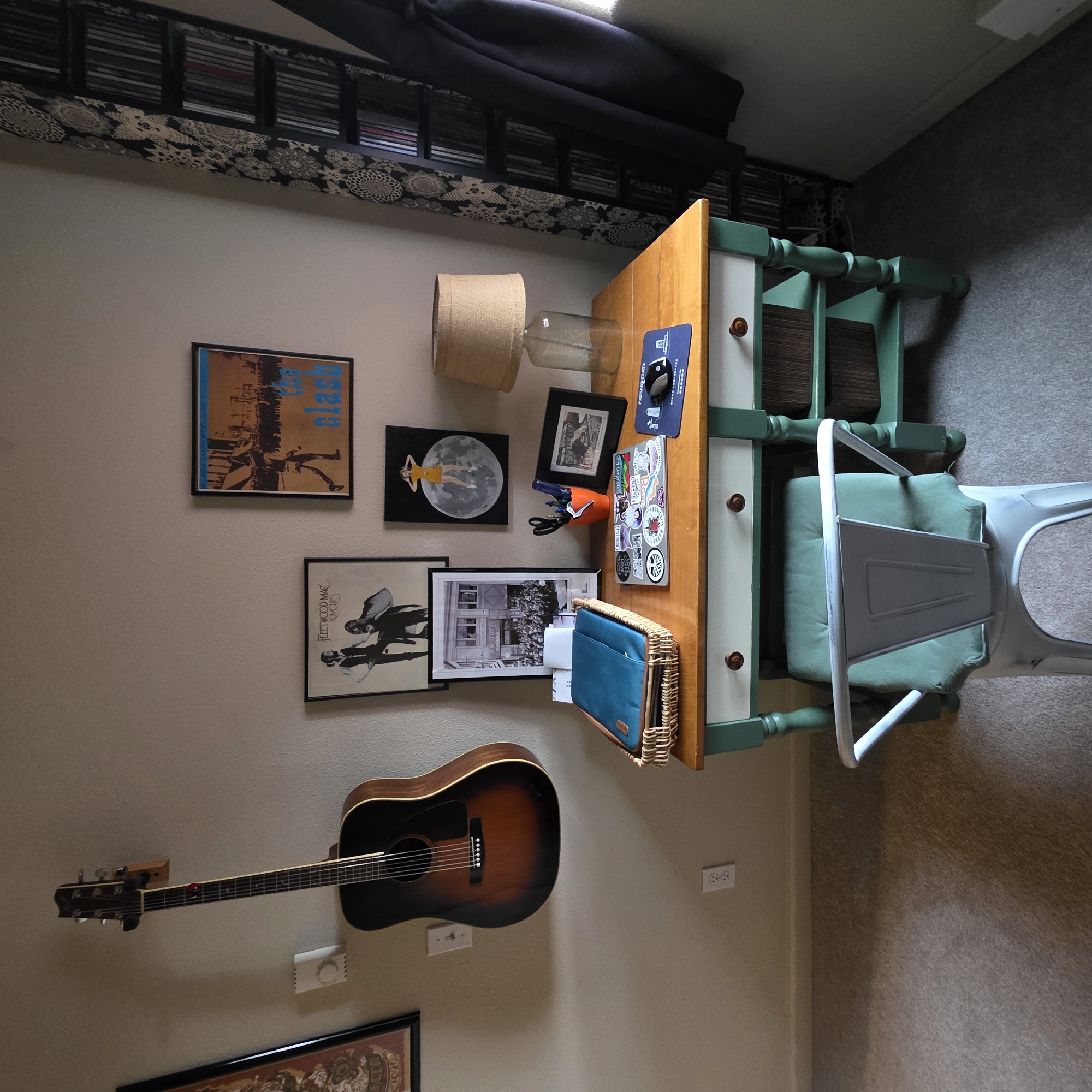 The image depicts an indoor scene with a desk positioned against a light-colored wall. On the wall, a dark-colored acoustic guitar is mounted horizontally. To the left of the guitar, a framed artwork with a circular design is partially visible. Below the guitar, a white electrical outlet is mounted on the wall.
The desk itself has a wooden top and a light-colored drawer front with dark wooden knobs. The base of the desk is painted green. On the desk, there is a woven basket containing a blue laptop case. A laptop covered in stickers sits in front of the basket. To the right of the laptop, an orange cup holds pens and scissors. A framed photograph is placed next to the cup, and a lamp with a burlap shade is situated on the far right of the desk. A blue mousepad with text and graphics is visible on the right side of the desk.
Below the desk, a green stool with a light green cushion is positioned in front of it. The desk has two shelves underneath, each holding a brown woven basket. The floor appears to be carpeted in a neutral tone.
To the right of the desk, a tall, black shelving unit filled with CDs or DVDs is visible. A dark-colored garment, possibly a coat, is hanging on the right side of the shelving unit. A patterned curtain with a black and white design is also present to the left of the shelving unit. Another framed artwork, a poster with text and figures, is hung on the wall above the desk, slightly to the left of center. A smaller framed artwork depicting a person standing on the moon is to the right of the larger poster.
The lighting in the room appears to be a mix of ambient light and light from the lamp on the desk. The overall scene suggests a home office or creative space. This scene is not located in Beaverton, United States of America.