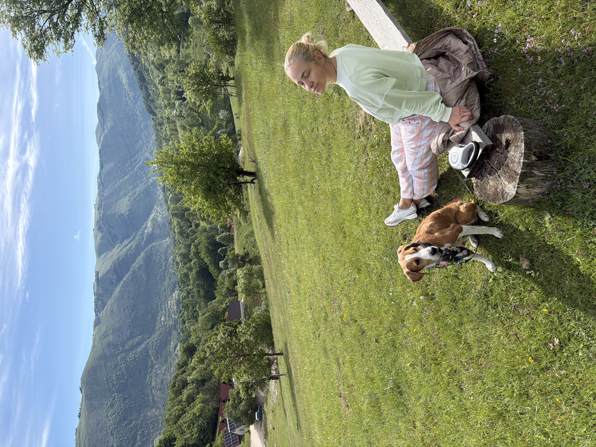 This image captures a tranquil outdoor scene featuring a woman and a dog on a grassy hillside, set against a stunning mountainous backdrop. A blonde woman, with her hair styled in a bun, is positioned towards the right of the frame. She wears a light green long-sleeved top, pink and white plaid pants, and white sneakers. She is looking downwards with a gentle expression, seemingly interacting with her dog or an object on the ground.
Beside her, a brown and white dog, possibly a beagle mix, lies comfortably on the vibrant green grass, holding a black and white object (likely a toy or the handle of its leash) in its mouth. A black and white retractable dog leash, along with a folded light brown jacket, rests on a rustic tree stump next to the woman. A wooden beam or bench is also partially visible, suggesting a resting spot.
The setting is a expansive, sun-drenched meadow filled with lush green grass and small wildflowers. In the mid-ground and background, majestic mountains covered with dense green forests stretch across the horizon, creating a breathtaking panoramic view. The sky above is clear and blue, adorned with soft, wispy white clouds. In the far distance on the mountain slopes, a few houses and what appear to be solar panels can be seen, indicating some human habitation within this natural landscape.
The bright daylight suggests it is either morning or afternoon, with the overall ambiance being calm and leisurely. The scene depicts a peaceful moment between the woman and her canine companion amidst the beautiful natural environment of Bijela, Montenegro. There is no visible text in the image.