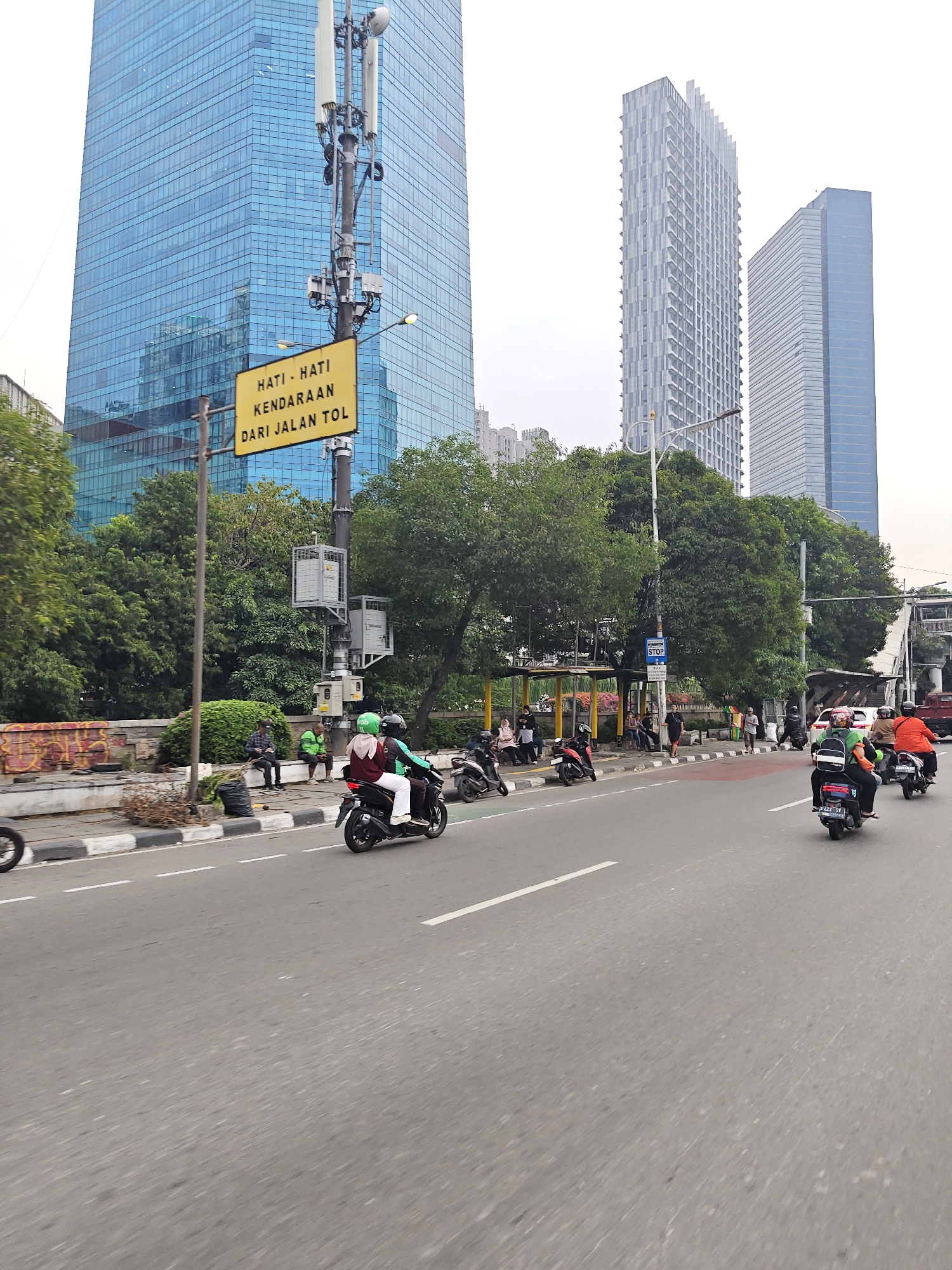 A street scene in West Jakarta, Indonesia, is captured with several high-rise buildings in the background. On the left, a tall, modern building with a blue glass facade dominates the skyline. To its right, a thinner, lighter-colored skyscraper stands, followed by another blue-tinted building. A prominent yellow sign with black text is affixed to a utility pole, reading "HATI - HATI KENDARAAN DARI JALAN TOL" (Be careful of vehicles from the toll road).
The foreground shows a busy street with multiple motorcycles and scooters. Several riders are visible, some wearing helmets, including green ones indicative of ride-sharing services. A few pedestrians are present on the sidewalk, seated or standing near a bus stop shelter. The road has clear lane markings, and a section of a raised curb with black and white stripes is visible. Trees and greenery line the side of the road, interspersed with sections of a decorative wall with graffiti. The sky is overcast, suggesting a cloudy day. The overall impression is a vibrant urban environment with ongoing traffic and pedestrian activity.