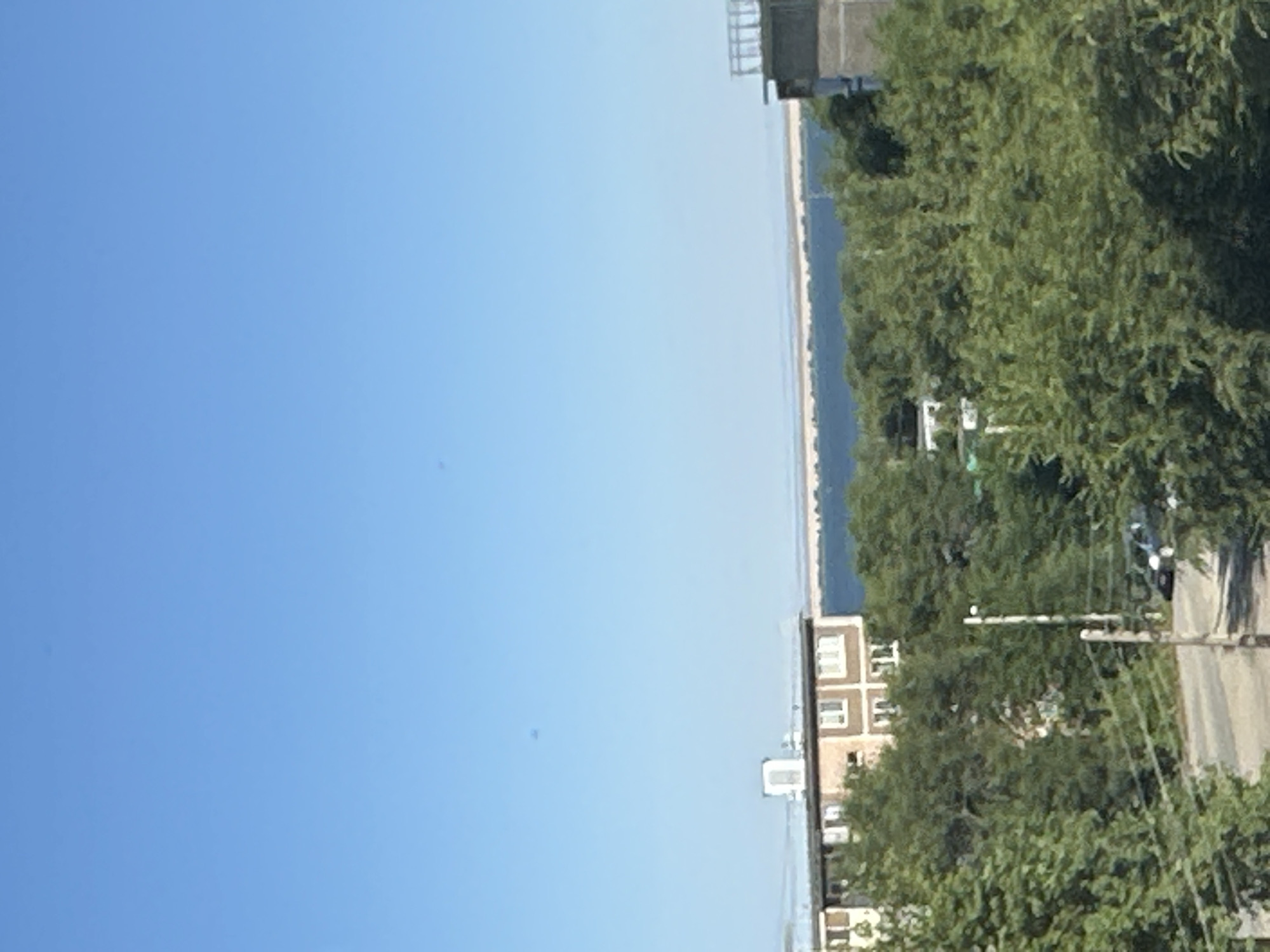 The image captures a cityscape under a clear blue sky, likely in Energetik, Russia, given the context. The foreground is dominated by lush green trees, obscuring much of the lower portion of the scene. A paved road is visible, with a car parked on the right side. A utility pole stands prominently near the road.
In the mid-ground, a multi-story building with a light-colored facade and dark trim is visible. It features multiple windows and appears to be an apartment complex or a similar residential structure. The building is partially surrounded by more trees.
Further in the background, a long, thin strip of land, possibly a shoreline or a natural barrier, is visible against a body of water. This body of water extends to the horizon, where it meets the sky. Another building, appearing more modern with a railing on top, is partially visible on the far right of the frame. The overall impression is a sunny day in a city with a significant presence of both urban structures and natural greenery.