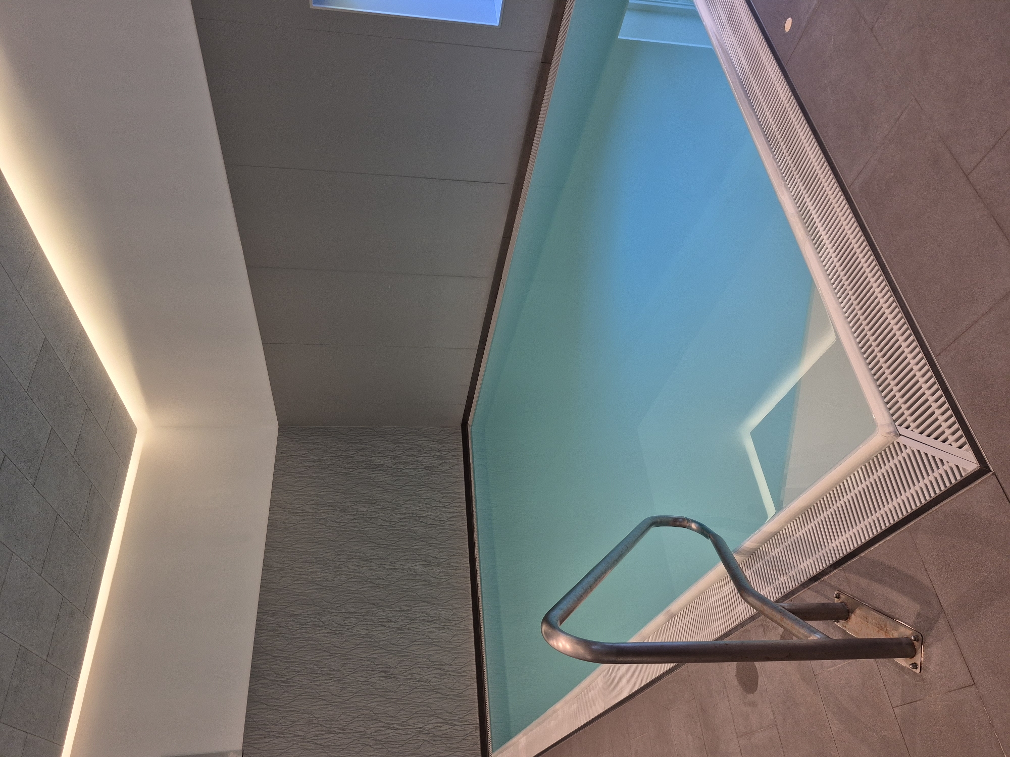 The image shows an indoor swimming pool with clear blue water. A stainless steel handrail is visible on the left side of the pool, leading down steps. The pool area is tiled with grey square tiles. The walls are a dark grey with a subtle textured pattern on the left and a smooth finish on the right. The ceiling is a light grey acoustic tile with indirect warm lighting illuminating the space from cove lighting. The scene appears to be a modern and well-maintained indoor swimming facility. The location is not explicitly stated in the image, so it cannot be determined to be in Schwäbisch Gmünd, Germany.