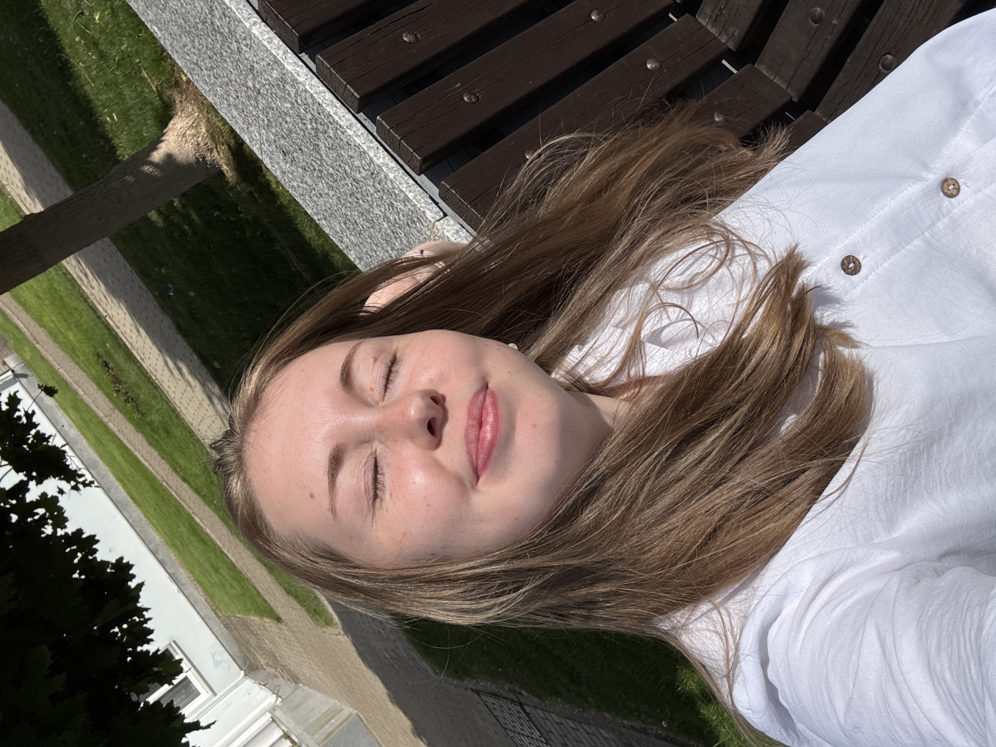 A single female individual is depicted in a high-angle, close-up outdoor self-portrait. She has fair skin with visible freckles on her nose and cheeks. Her eyes are closed, and her lips are slightly parted in a gentle smile. Her long, light brown hair is parted on the left and falls over her shoulders. She wears a white collared shirt featuring two visible light brown, circular buttons with a textured, natural appearance. A small, spherical, light-colored earring is visible in her left earlobe. Her right arm or hand is partially visible behind her head.
The individual is seated on a dark brown wooden bench with horizontal slats and visible metallic fastenings. The bench has a top edge made of light grey, speckled stone, consistent with granite. The background shows an outdoor public space under strong sunlight, casting defined shadows. The ground features parallel strips of light grey-beige paving and green grass. A tree trunk is visible in the mid-right background, and the dark green foliage of another tree partially overhangs the top left corner. A light-colored building, likely white or very pale grey, with a visible window, stands in the upper left background. The specific city and country of this location are unknown.