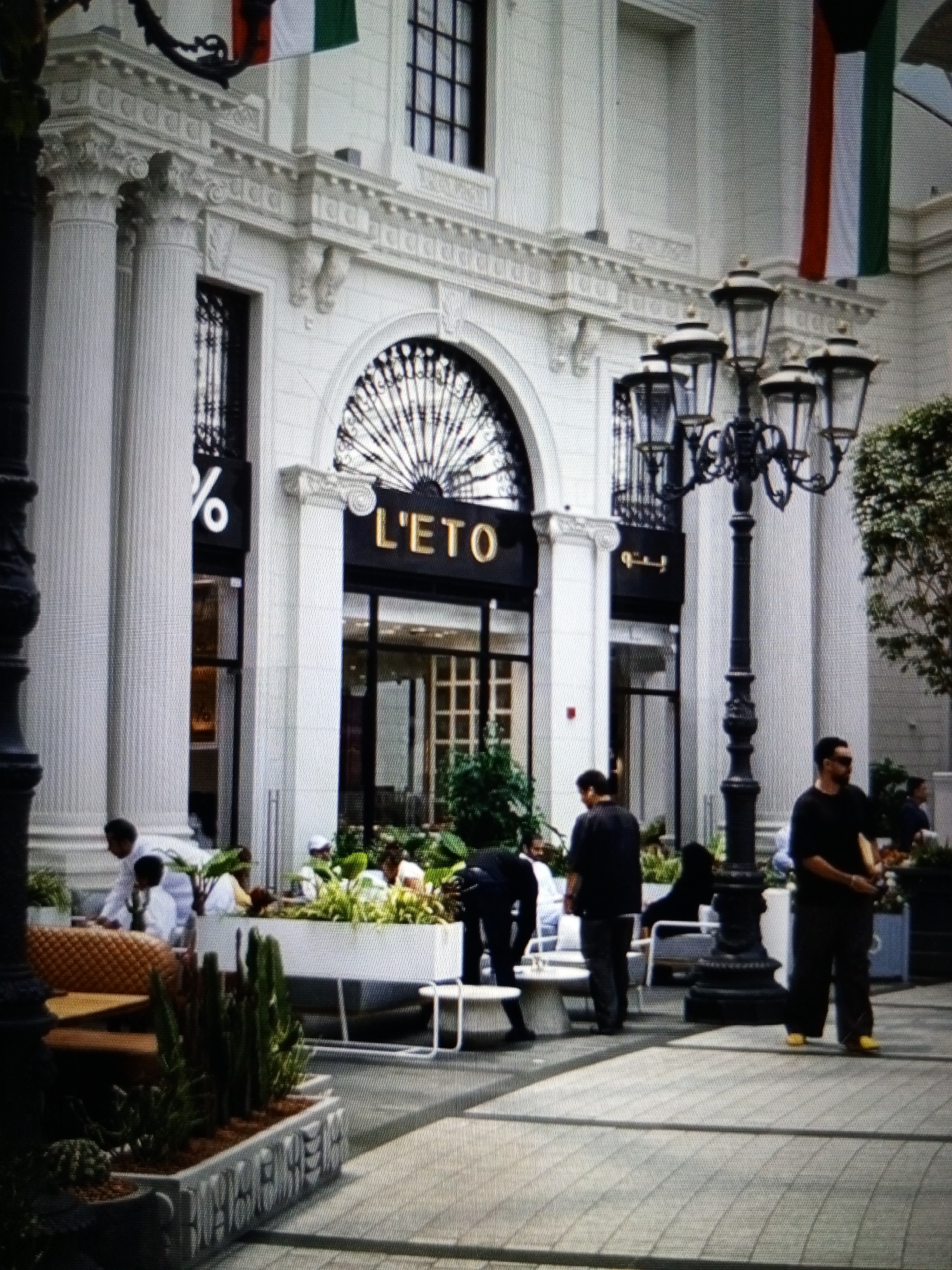 A medium shot captures the exterior of a building with classical architectural elements, including large white columns, ornate moldings, and a decorative arched window. Two national flags, red, white, and green, are draped on the facade. In front of the building, a group of people are visible. To the left, near the entrance with a sign displaying "%", a person in white is interacting with another person. Further to the right, a lamppost with multiple lights stands prominently. Several individuals are standing and walking on the paved area. One man, wearing a black t-shirt and dark pants with yellow shoes, is standing in the foreground, looking down. Another man in a black t-shirt and dark pants is standing to his left. A planter with various green plants is situated in front of a white seating area. The scene appears to be a public outdoor space, possibly a commercial area or a plaza. The location is identified as Golbembal, Nigeria.